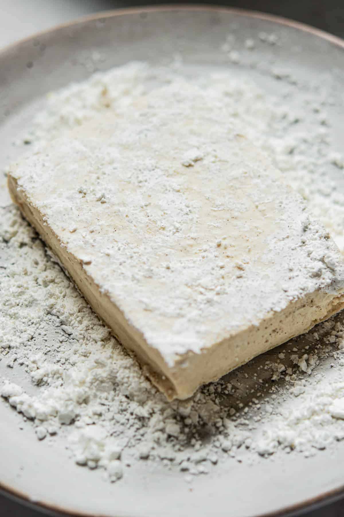 A rectangular slab of marshmallow dusted with powdered sugar sits on a plate, with more powdered sugar scattered around.