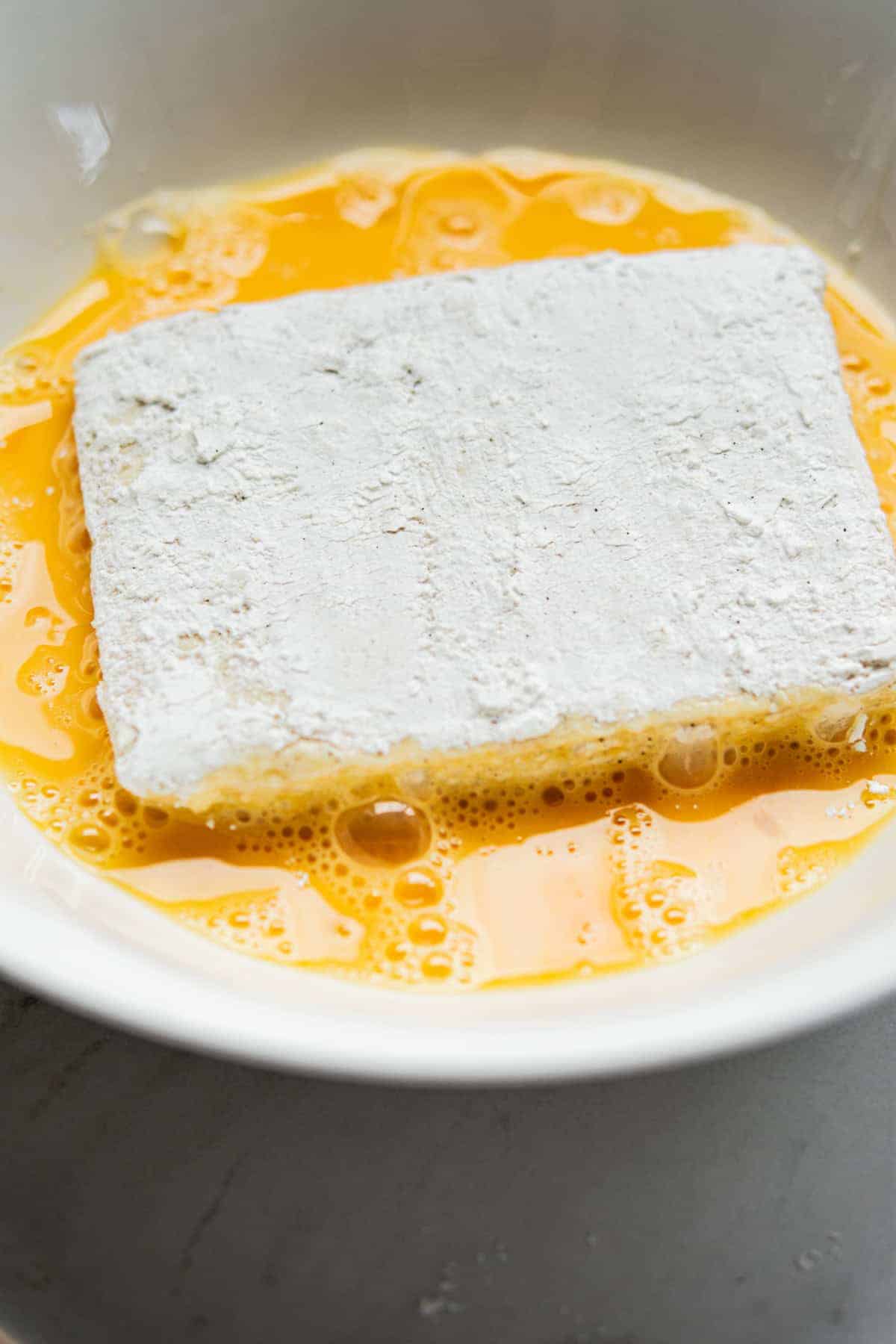 A rectangular slice of tofu coated in flour sits in a bowl of beaten eggs, ready for breading or frying.
