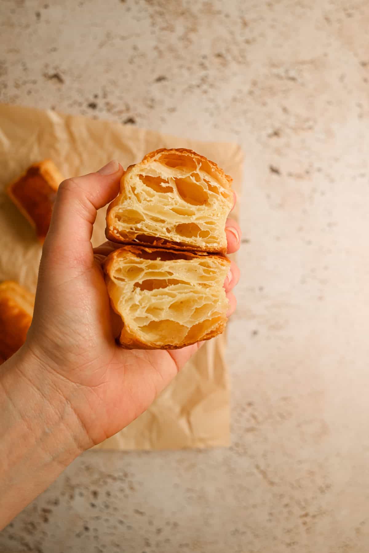 A hand holds a flaky, golden pastry broken in half, showing its airy, layered interior. The background is a neutral, textured surface with more pastries on brown parchment paper.
