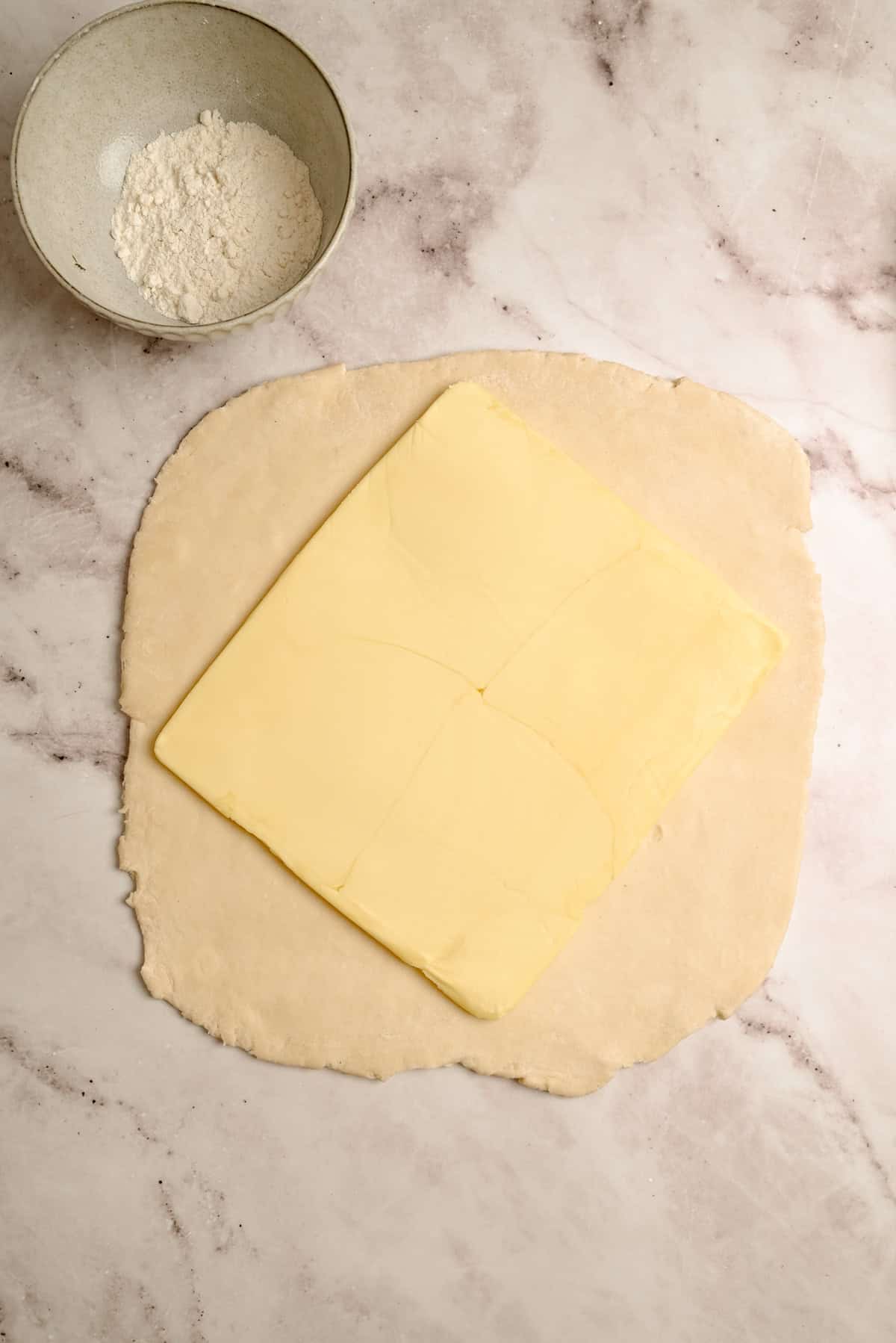 A slab of butter is placed on a sheet of rolled-out dough on a marble surface, with a bowl of flour nearby.
