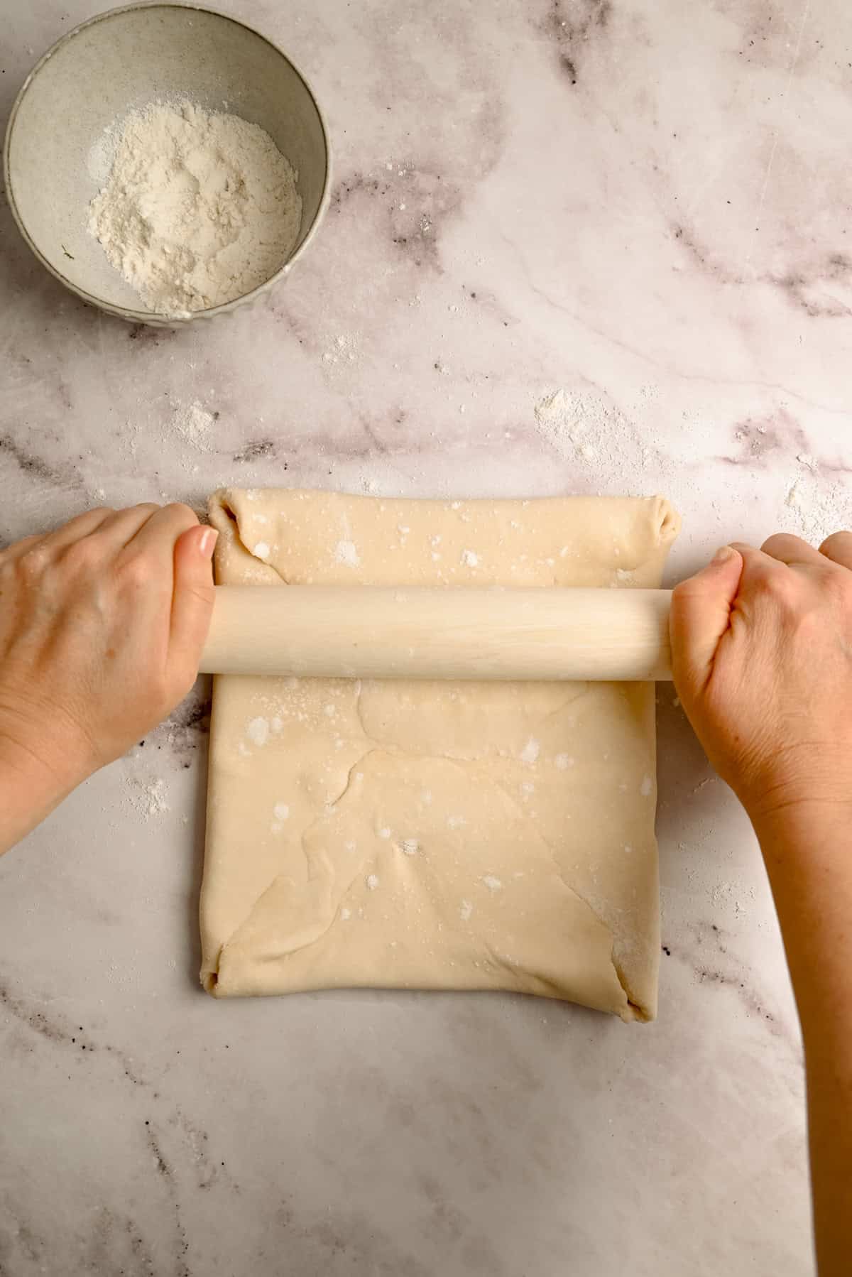 Hands rolling out folded dough with a rolling pin on a floured marble surface, with a bowl of flour nearby.