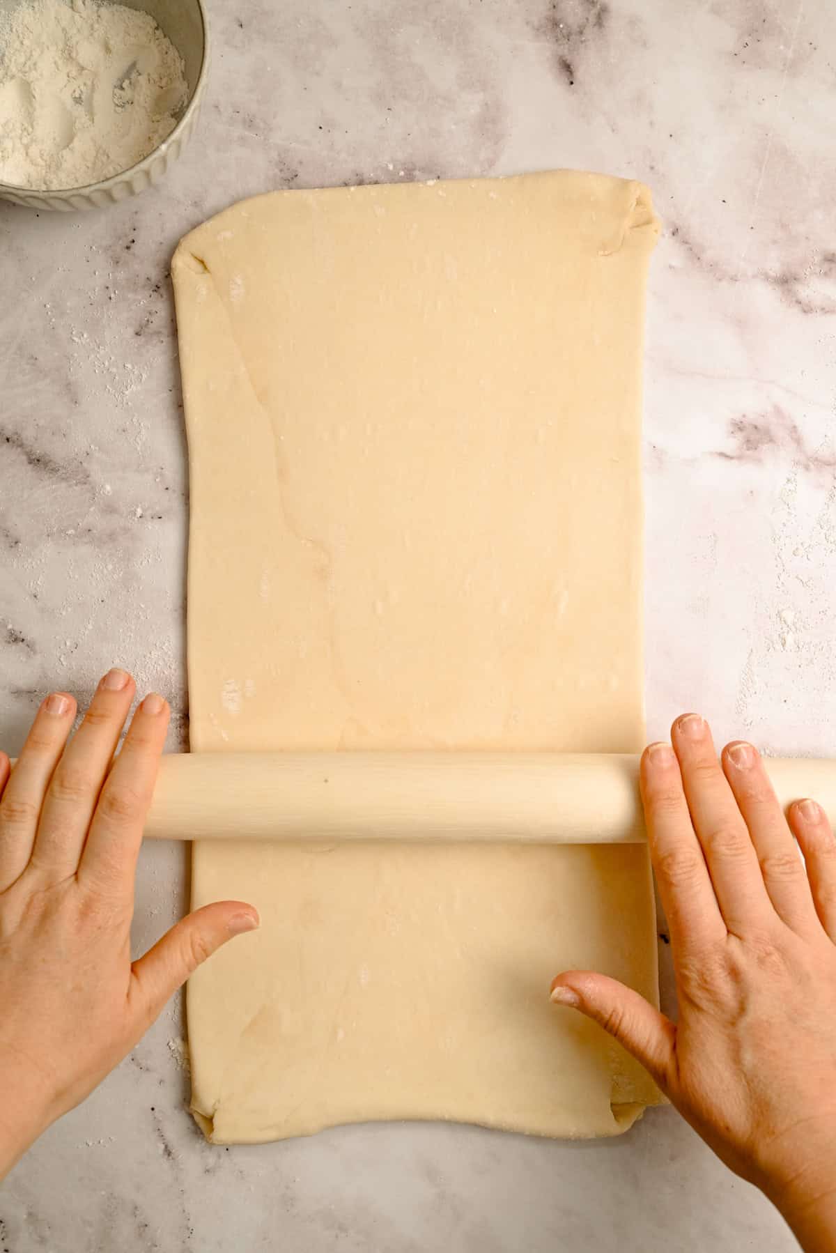 A pair of hands uses a rolling pin to flatten a rectangular sheet of dough on a lightly floured, marble-like surface. A bowl of flour sits in the upper left corner.