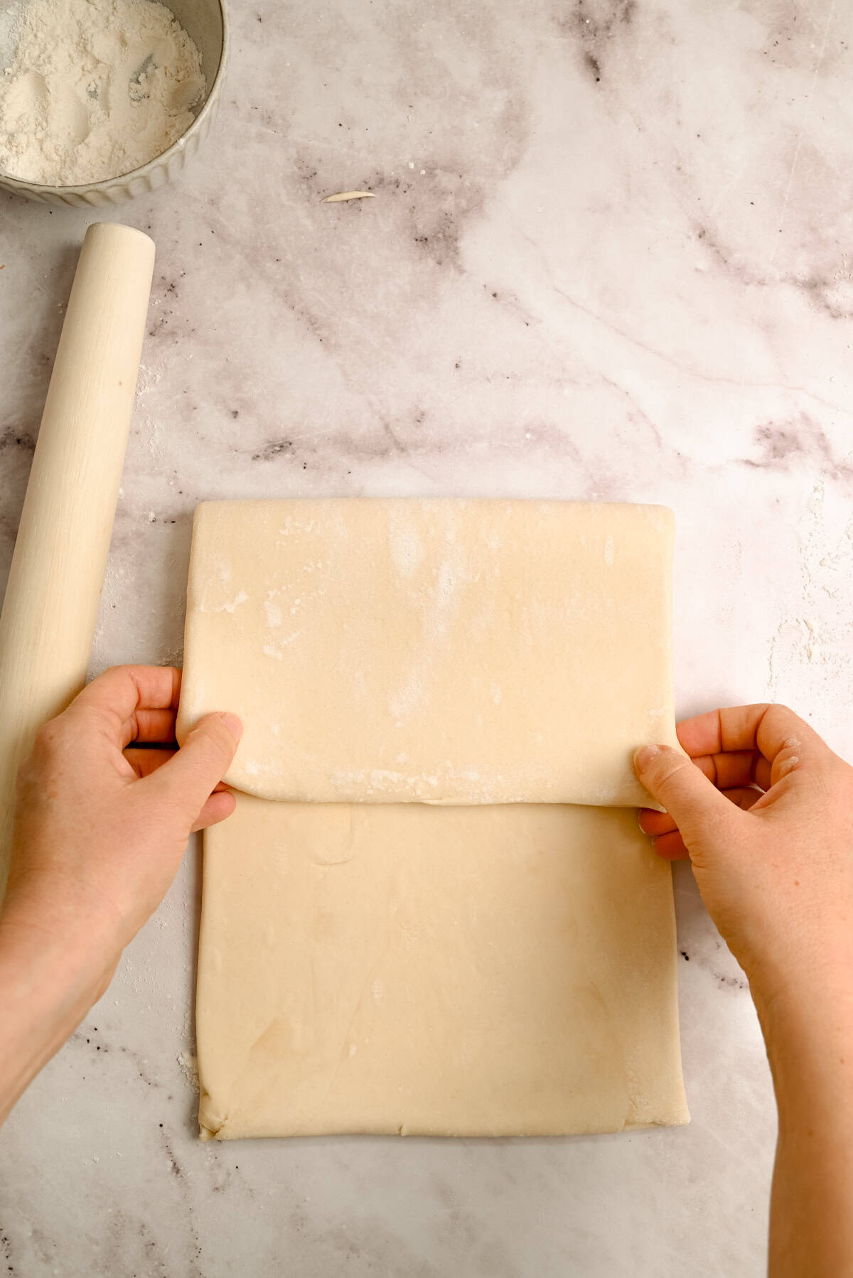 Hands folding a sheet of dough on a floured marble surface, with a rolling pin and a bowl of flour nearby.