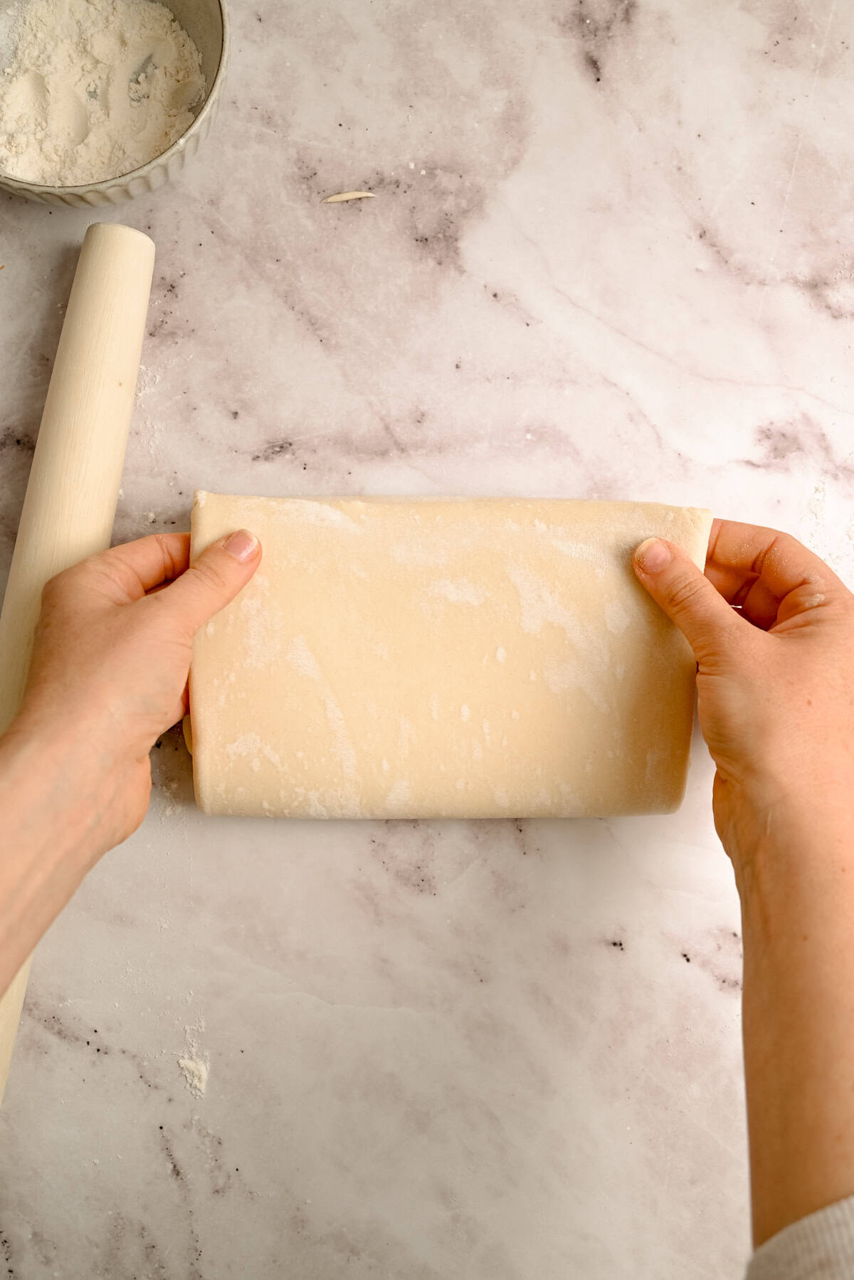 A pair of hands folding a sheet of dough on a floured surface, with a rolling pin and a bowl of flour nearby.