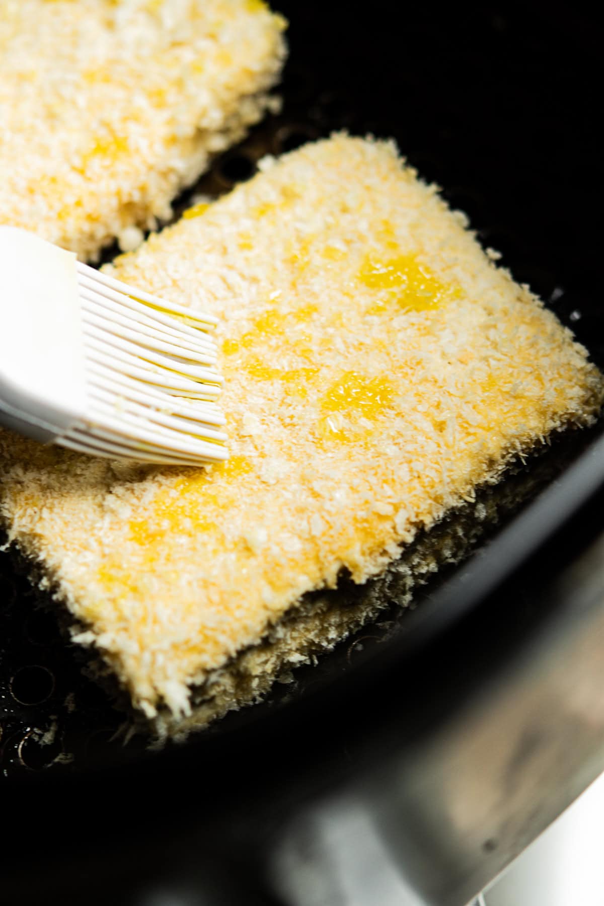 A close-up of a breaded tofu item being brushed with oil using a white silicone brush, placed inside an air fryer basket.