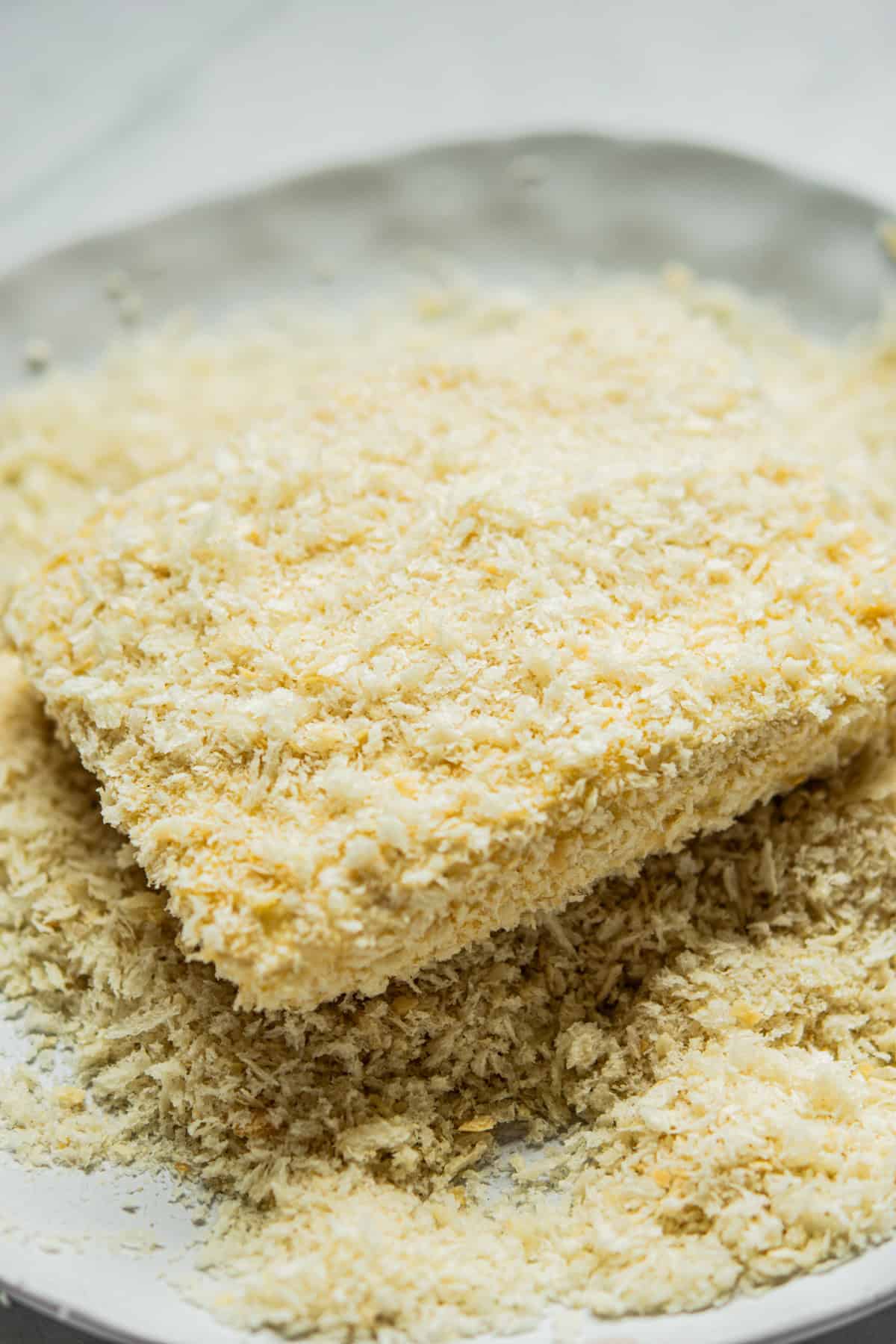 A close-up of a rectangular piece of tofu coated in panko bread crumbs on a white plate, with loose crumbs scattered around it.
