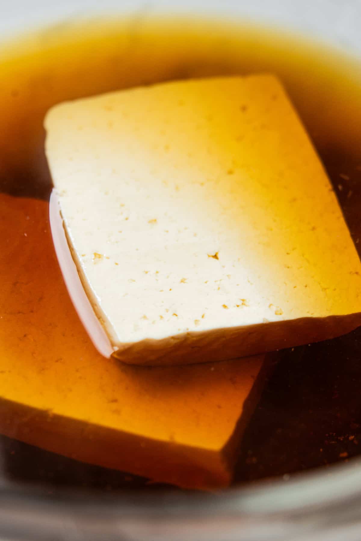 Close-up of two tofu slices marinating in a dark brown liquid, likely soy sauce, inside a glass container. The tofu appears to be absorbing the marinade.