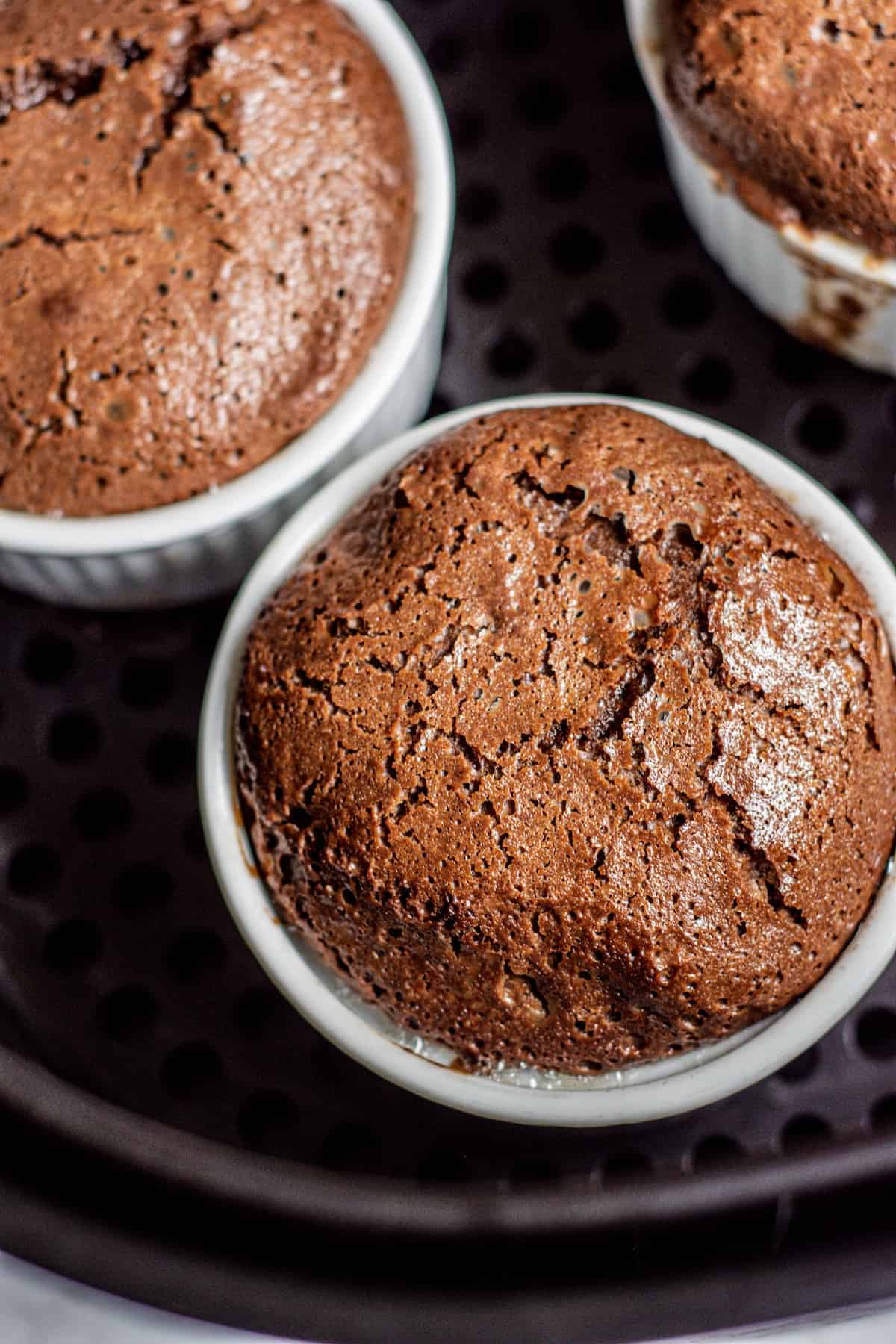 Close-up of individual chocolate soufflés in white ramekins, with cracked, risen tops and a rich, moist texture, sitting on a dark surface.