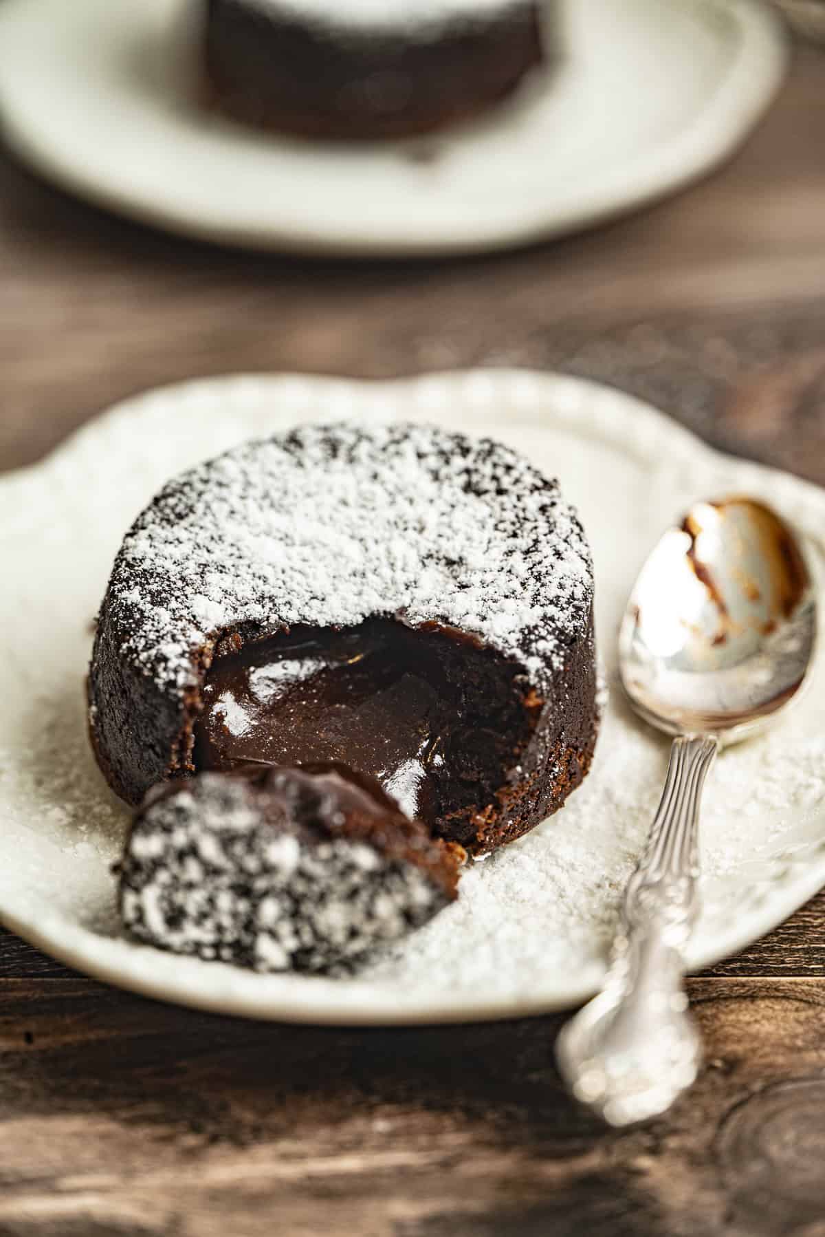 An air fryer chocolate lava cake dusted with powdered sugar sits on a plate. A spoon and a piece of the cake reveal the gooey chocolate center.