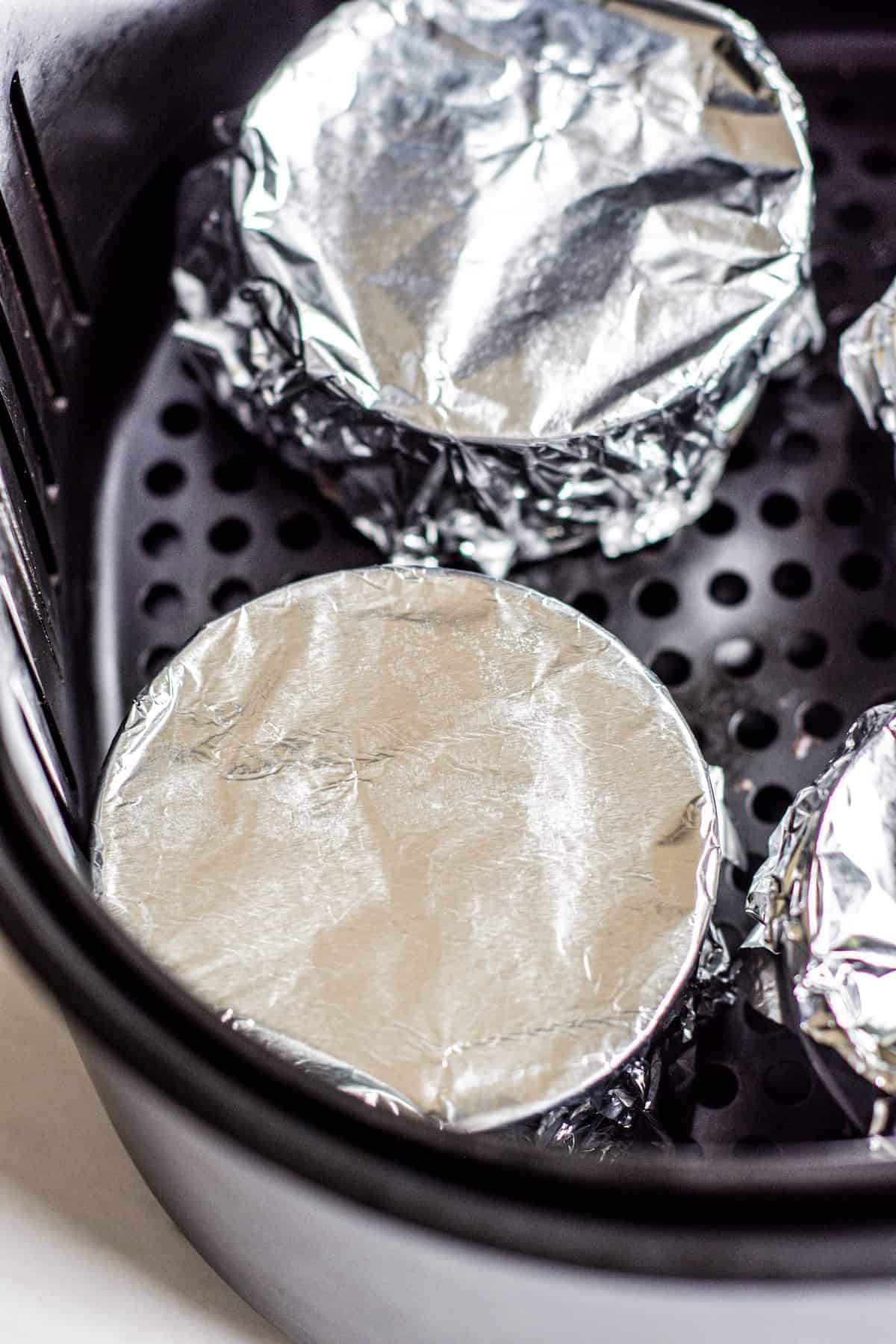 Four round dishes covered with aluminum foil sit inside an air fryer basket, ready for cooking.