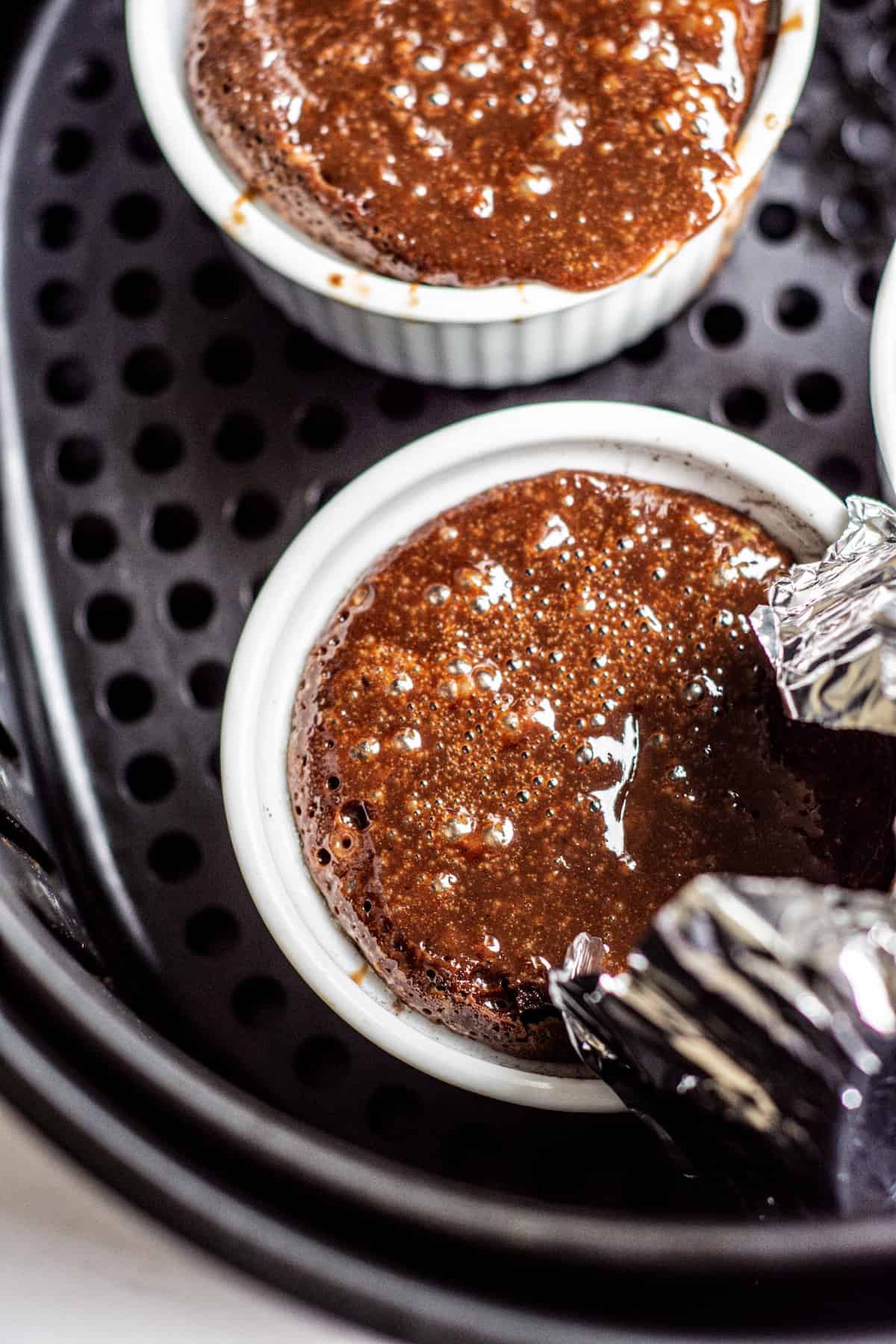 Two white ramekins filled with chocolate lava cake batter inside an air fryer basket, with one ramekin partially covered by a piece of aluminum foil.