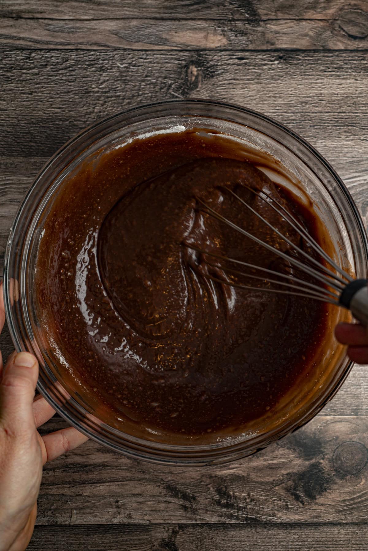 A hand holding a glass bowl of thick chocolate batter while whisking.