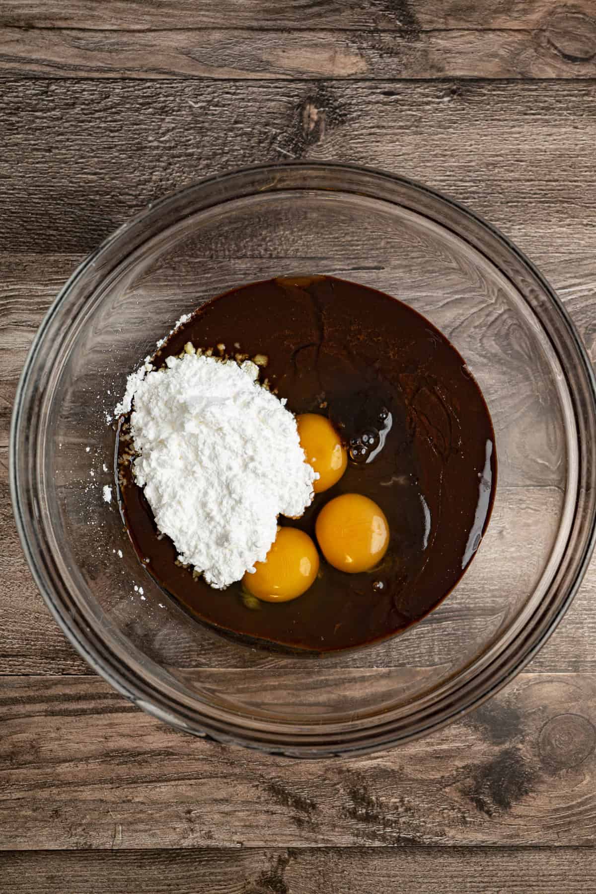 A glass mixing bowl on a wooden surface holds eggs, white powdered sugar, and chocolate batter.