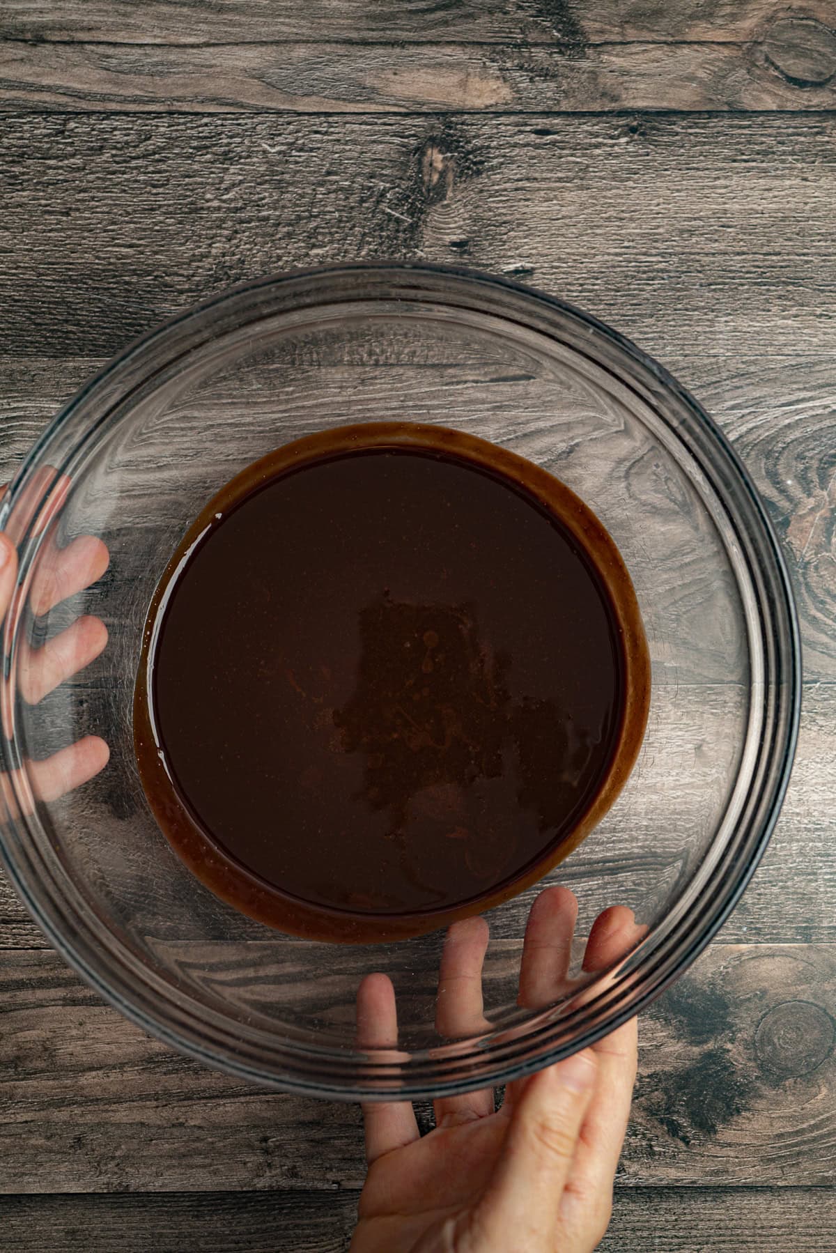 A person holds a clear glass bowl filled with smooth, melted chocolate on a wooden surface.