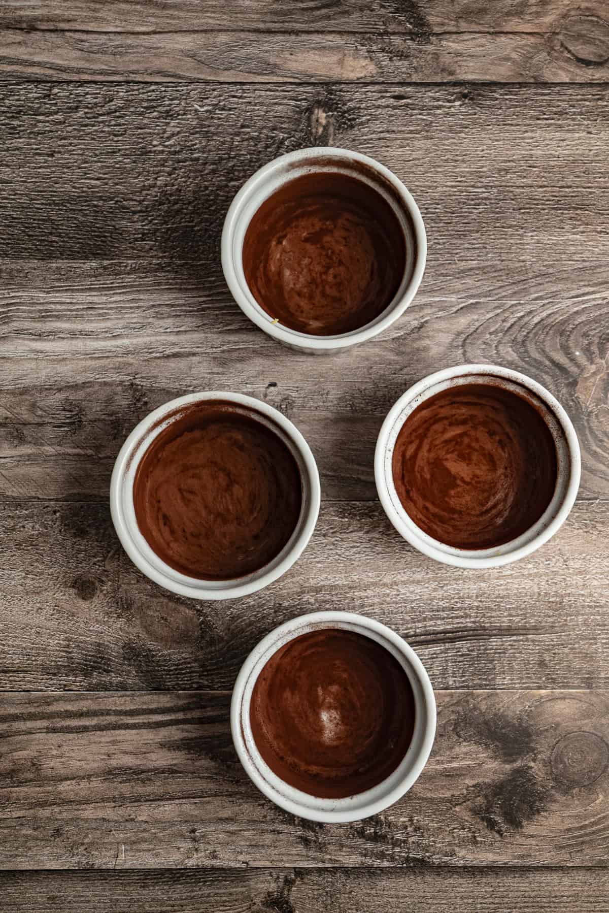 Four white ramekins with a thin layer of chocolate inside sit on a rustic wooden surface.