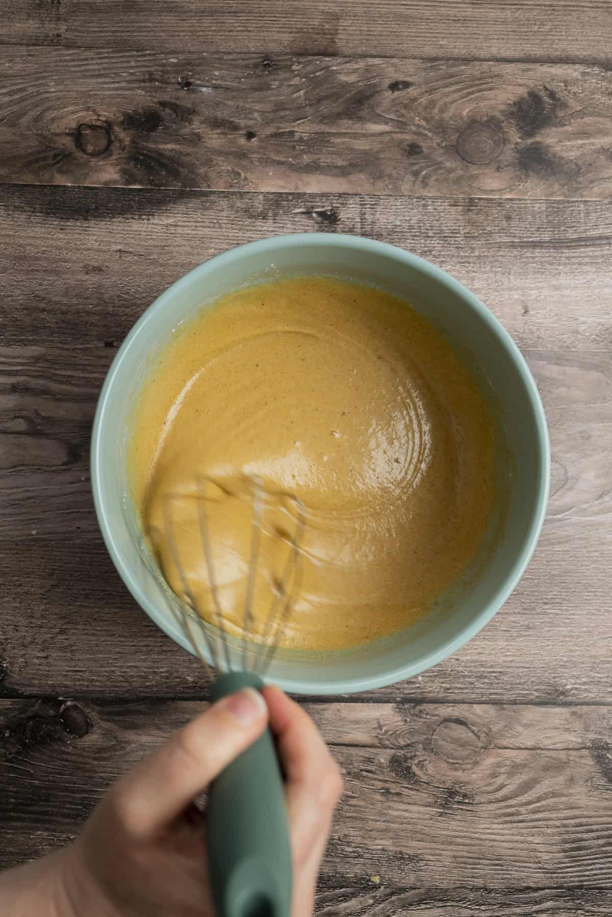 A hand holding a whisk is mixing yellow dough in a light blue bowl placed on a wooden surface.