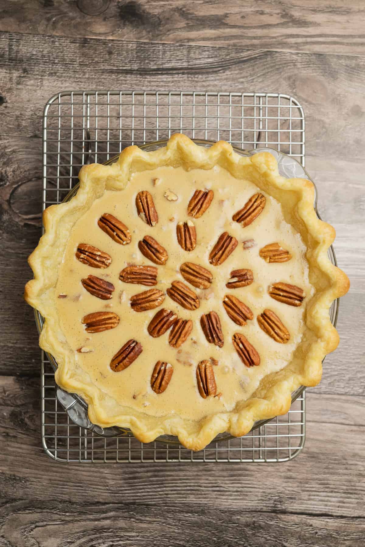 A freshly baked pecan pie with a golden curly crust sits on a cooling rack over a wooden surface. Whole pecans are arranged in a circle on top of the filling.