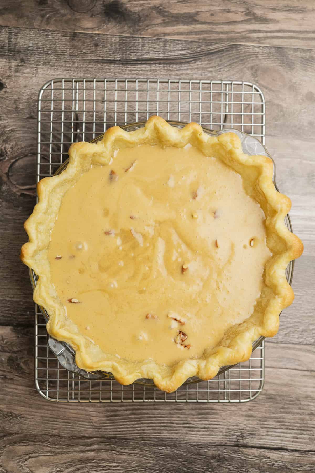 A freshly baked pie with a golden curly crust and a smooth light brown filling sits on a wire cooling rack over a wooden surface. You can see small pieces in the filling.