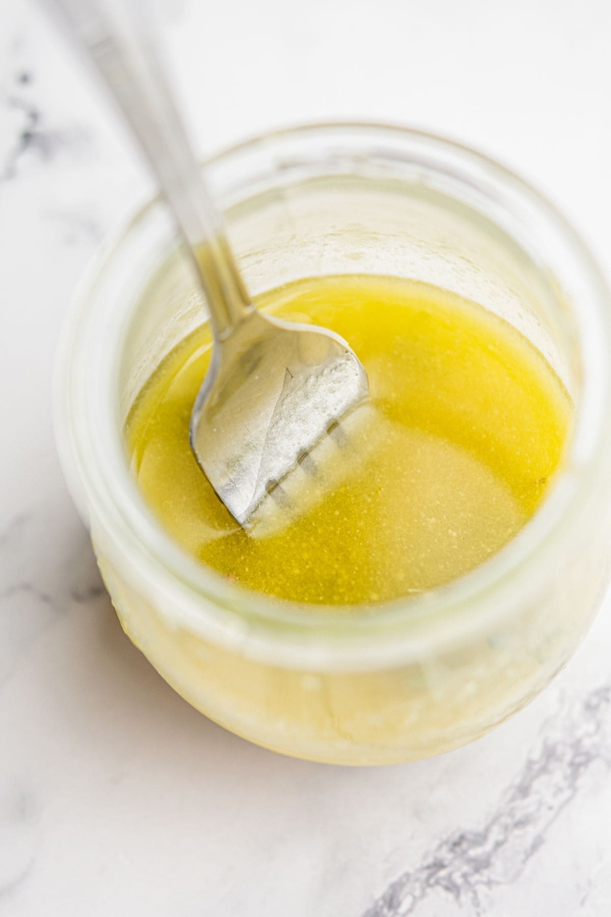 A close-up of a fork in a glass jar filled with melted yellow butter, placed on a white marble surface.