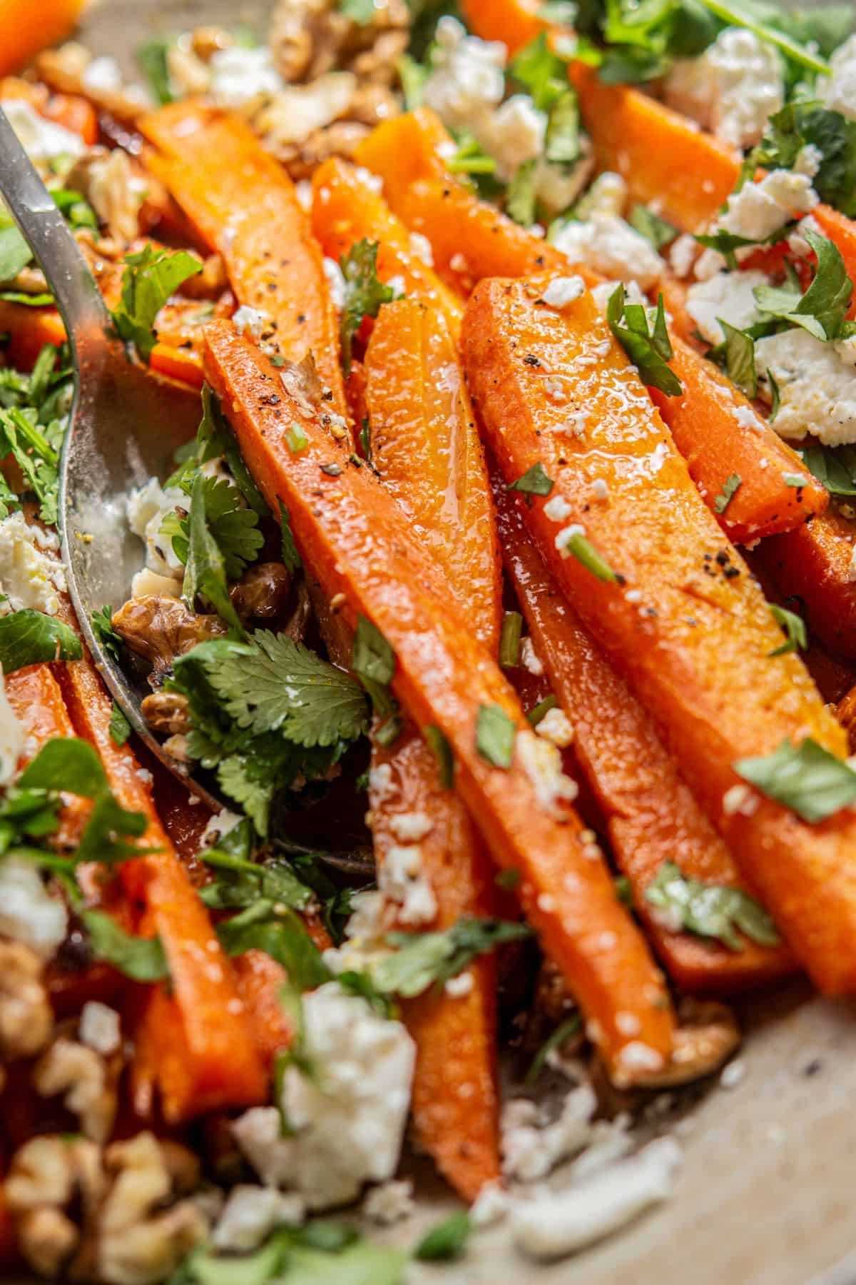 Close-up of roasted carrot sticks topped with crumbled feta cheese, chopped cilantro, and walnuts. A metal spoon is partially visible among the carrots, emphasizing the dish’s texture and vibrant colors.