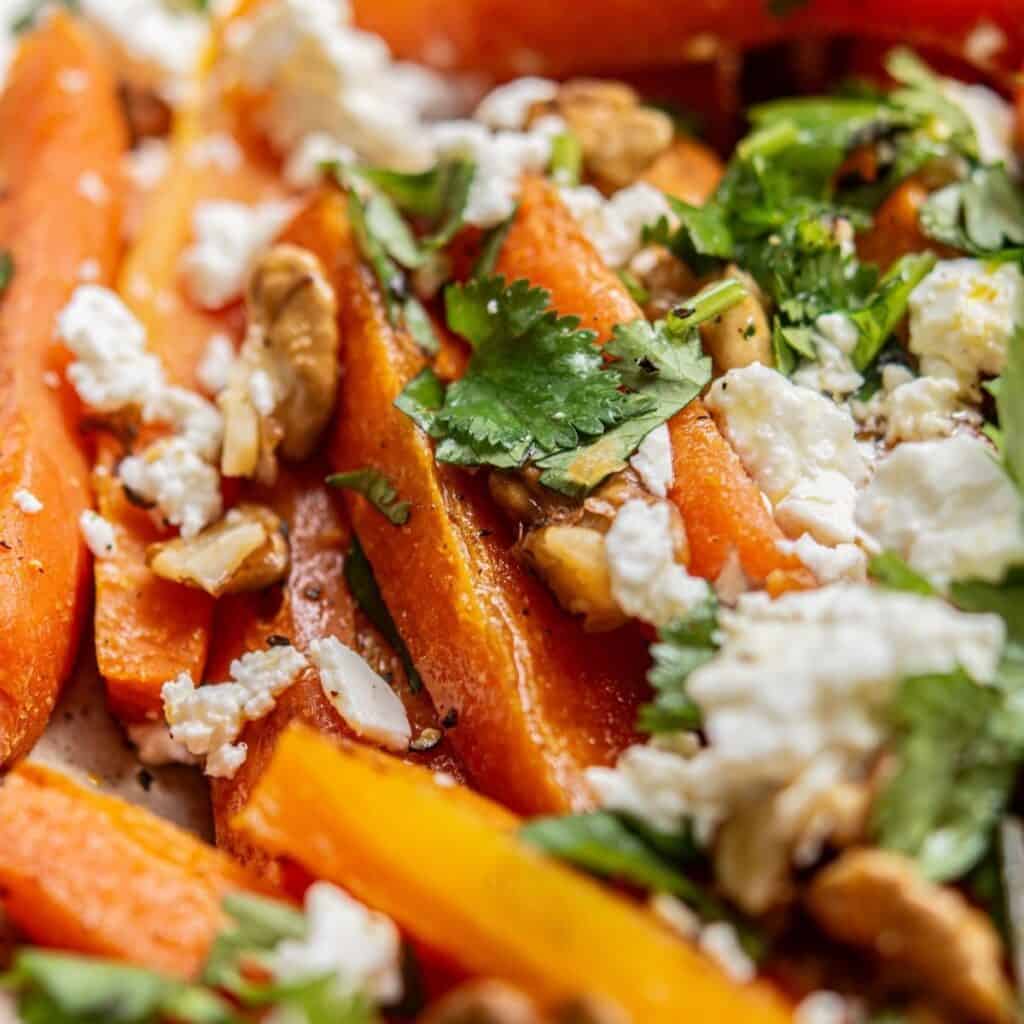 Close-up of roasted carrot sticks topped with crumbled feta cheese, chopped walnuts, and fresh cilantro leaves. The vibrant colors and textures of the ingredients are clearly visible.