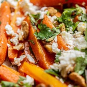 Close-up of roasted carrot sticks topped with crumbled feta cheese, chopped walnuts, and fresh cilantro leaves. The vibrant colors and textures of the ingredients are clearly visible.