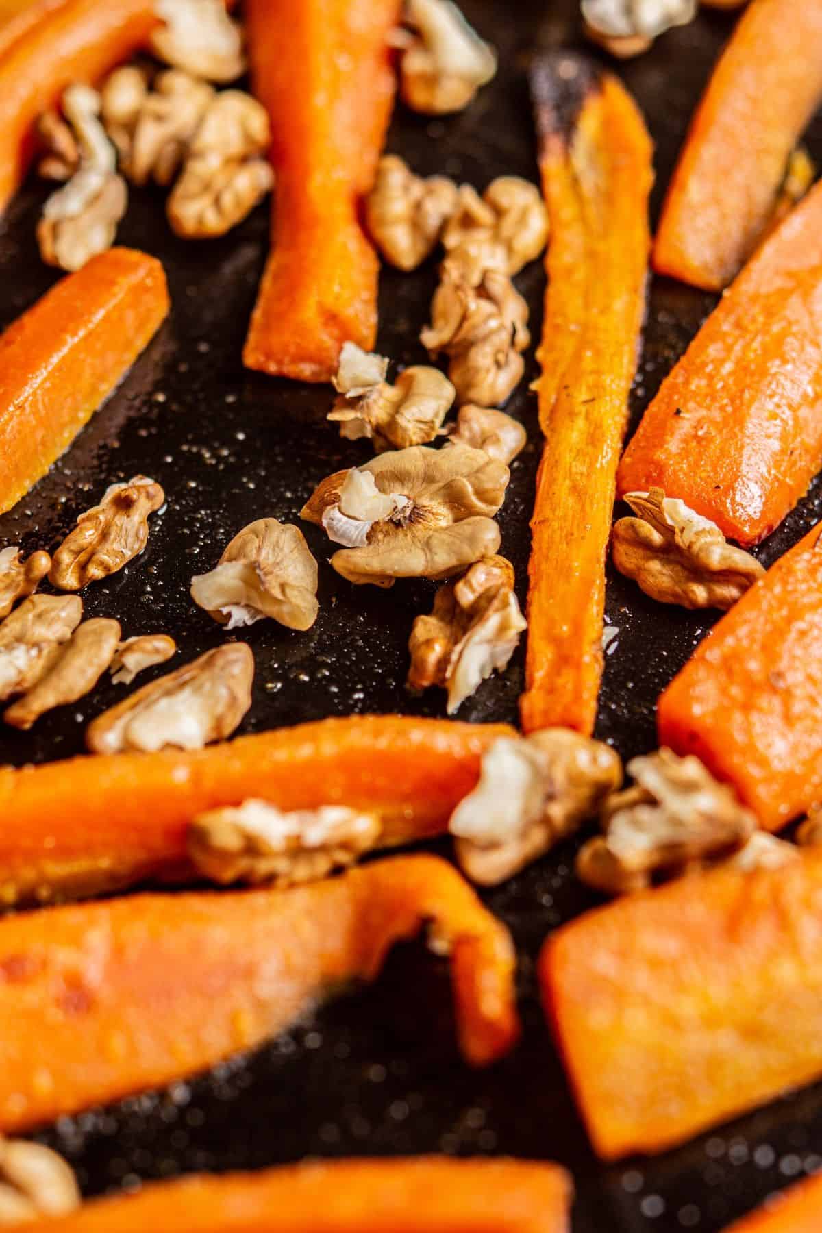 Close-up of roasted carrot sticks and walnut pieces on a dark baking sheet. The carrots are slightly caramelized, and the walnuts are scattered among them, both appearing lightly toasted.