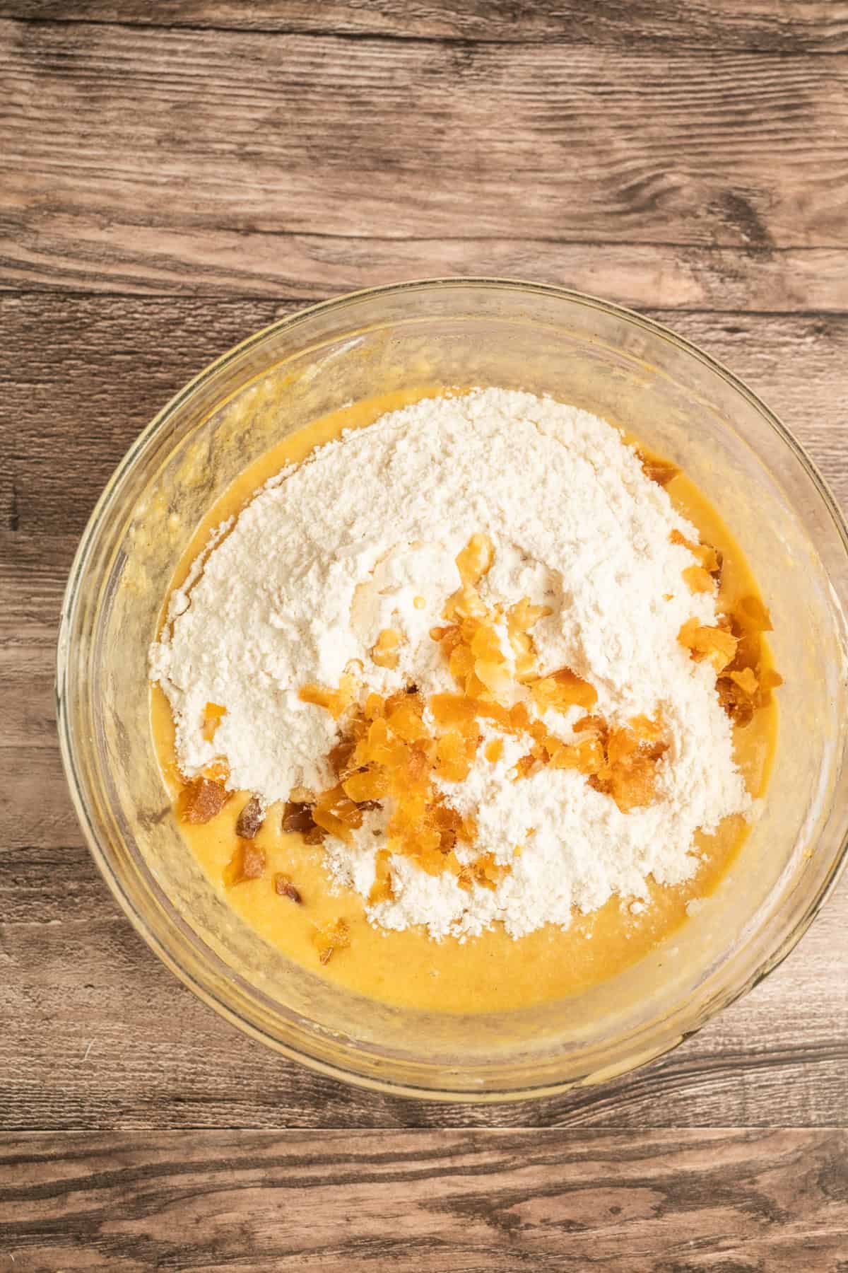 A glass bowl containing a baking mixture with flour and small pieces of dried fruit on top, resting on a wooden surface.