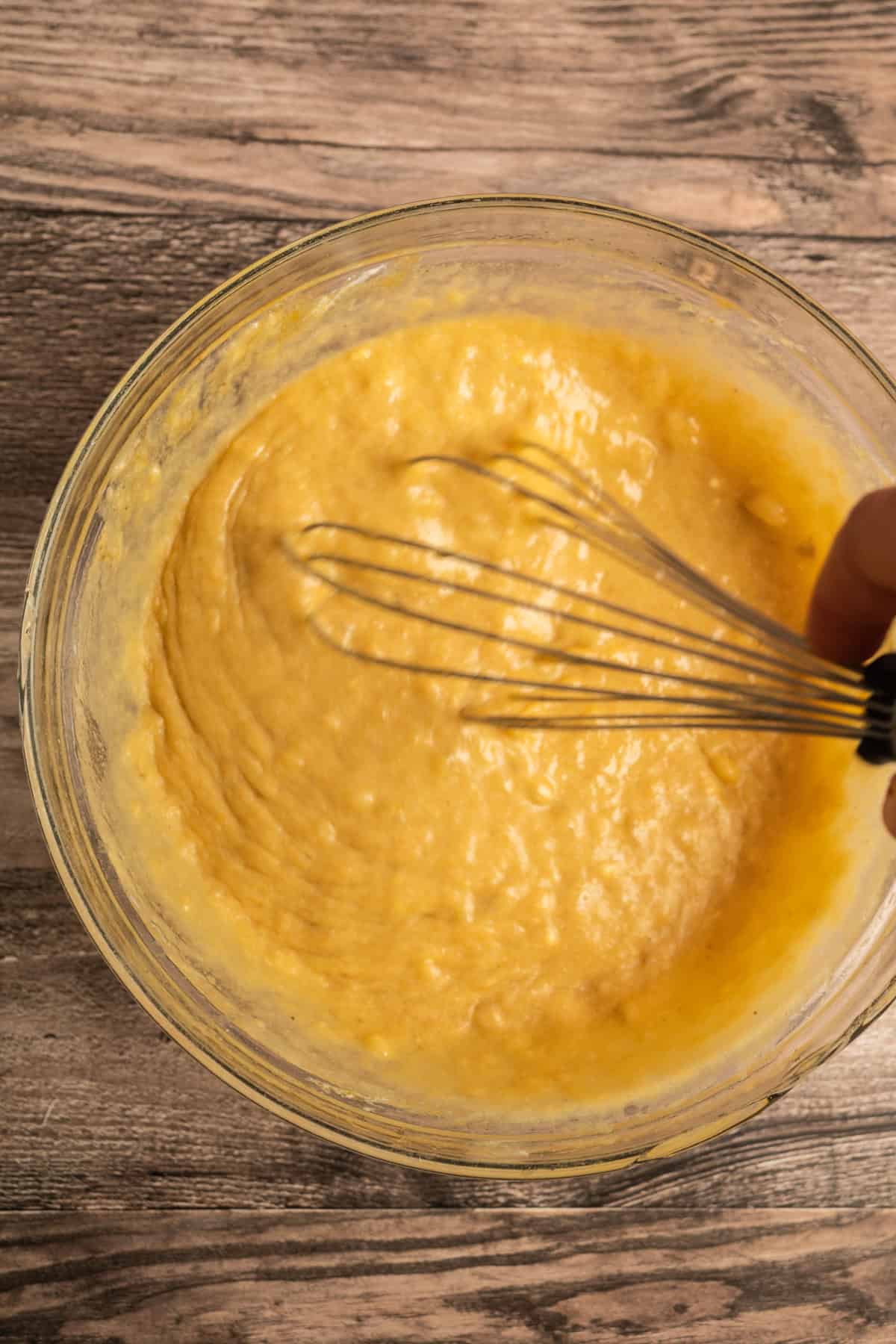 A hand holds a whisk, mixing thick yellow batter in a glass bowl on a wooden surface.