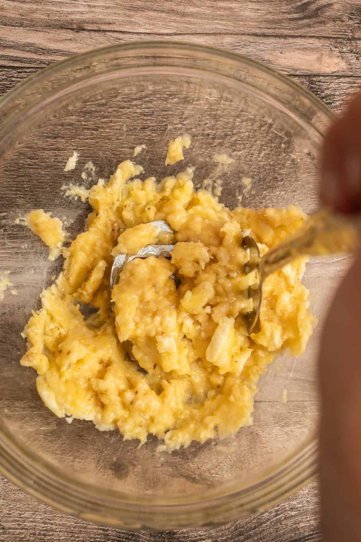 A hand uses a fork to mash ripe bananas in a clear glass bowl on a wooden surface.