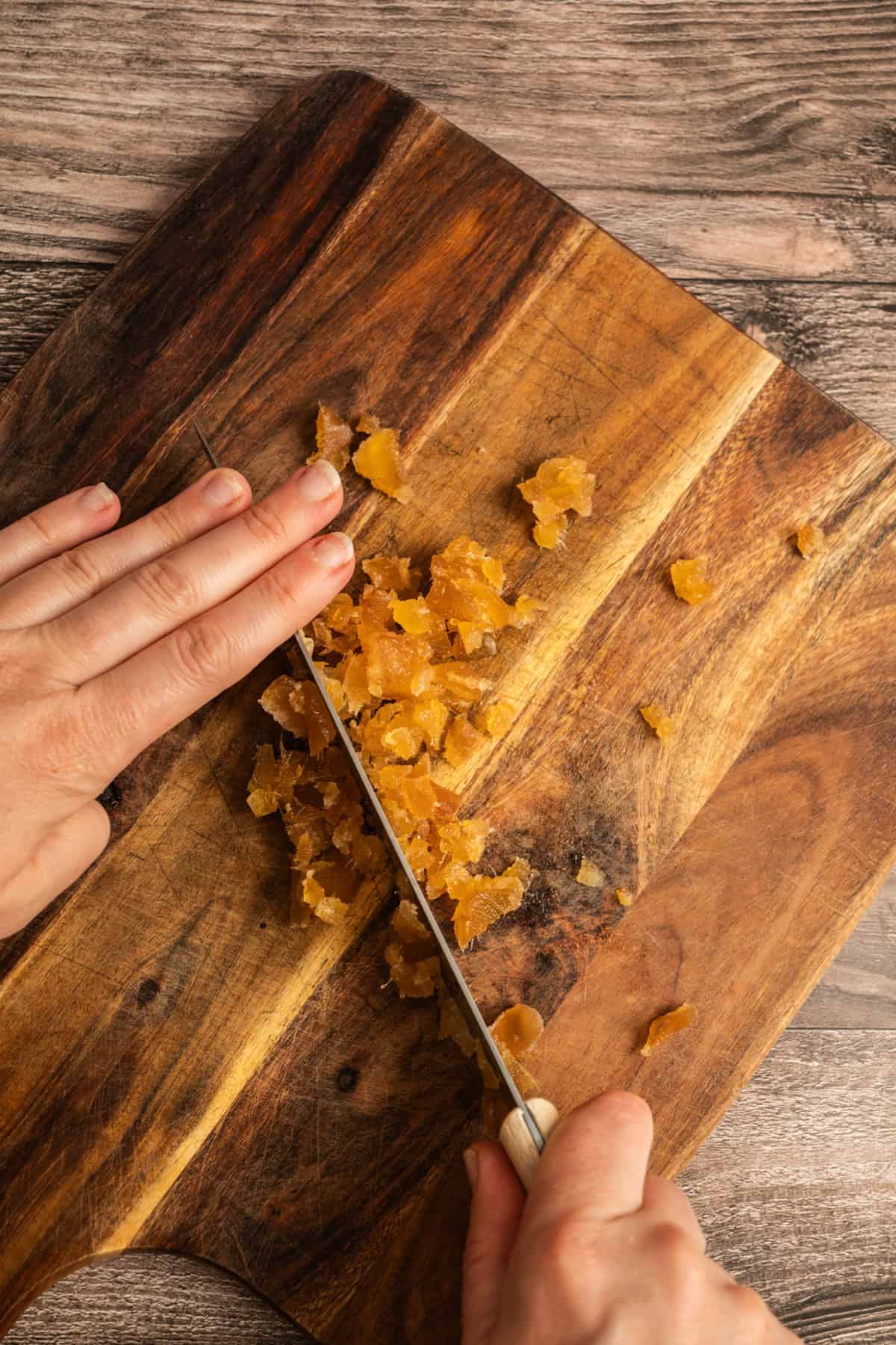 A person finely chops pieces of crystallized ginger on a wooden cutting board with a small knife.