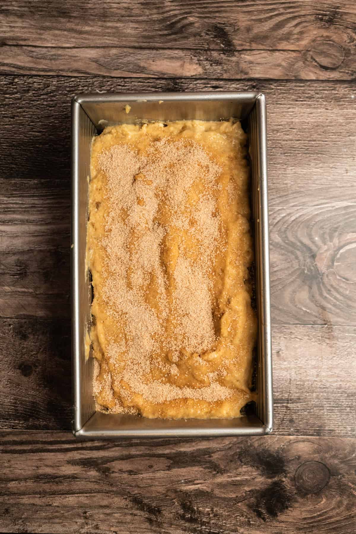 A rectangular metal baking pan filled with unbaked yellow batter, topped with a layer of cinnamon sugar, sits on a wooden surface.