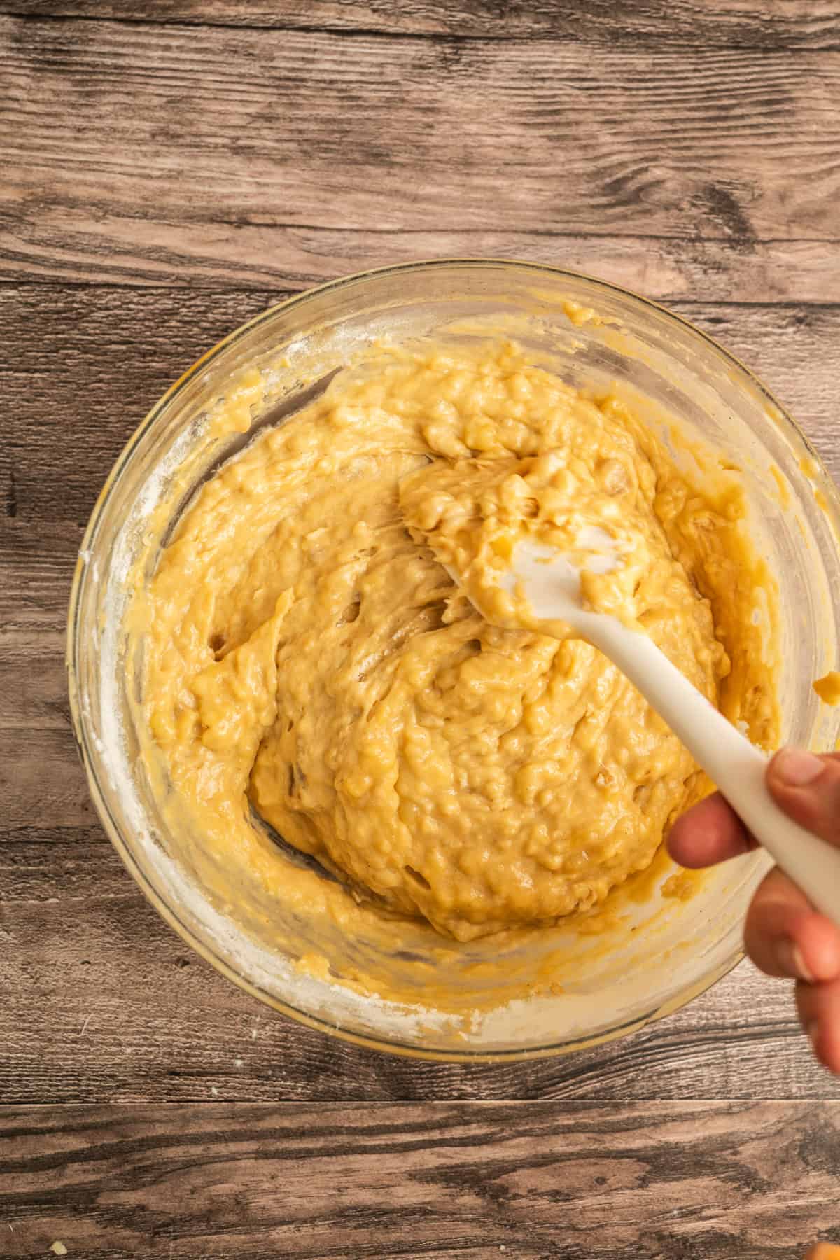 A hand holds a white spatula, mixing thick, yellowish batter in a glass bowl placed on a wooden surface.