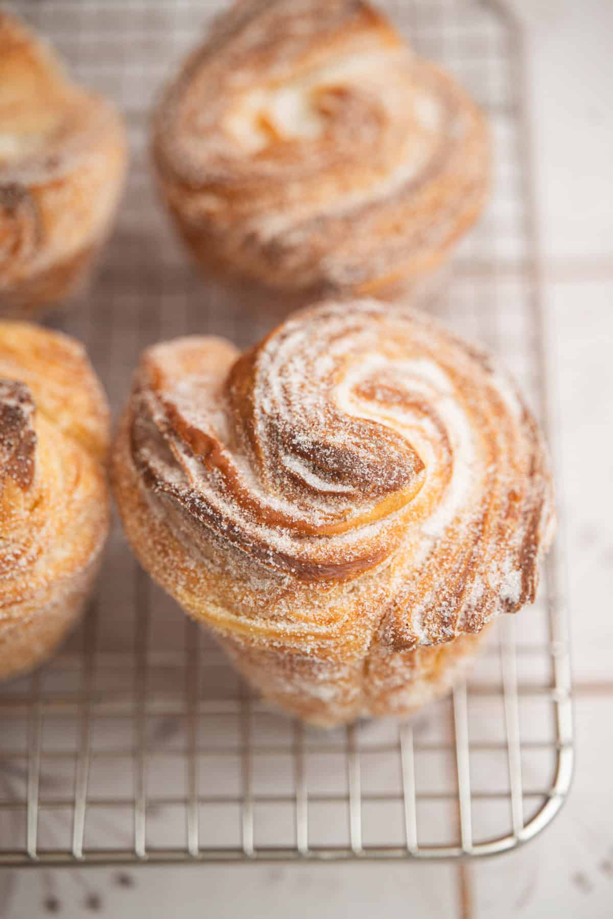 A close-up of a golden, swirled pastry dusted with sugar, resting on a metal cooling rack with similar pastries in the background.