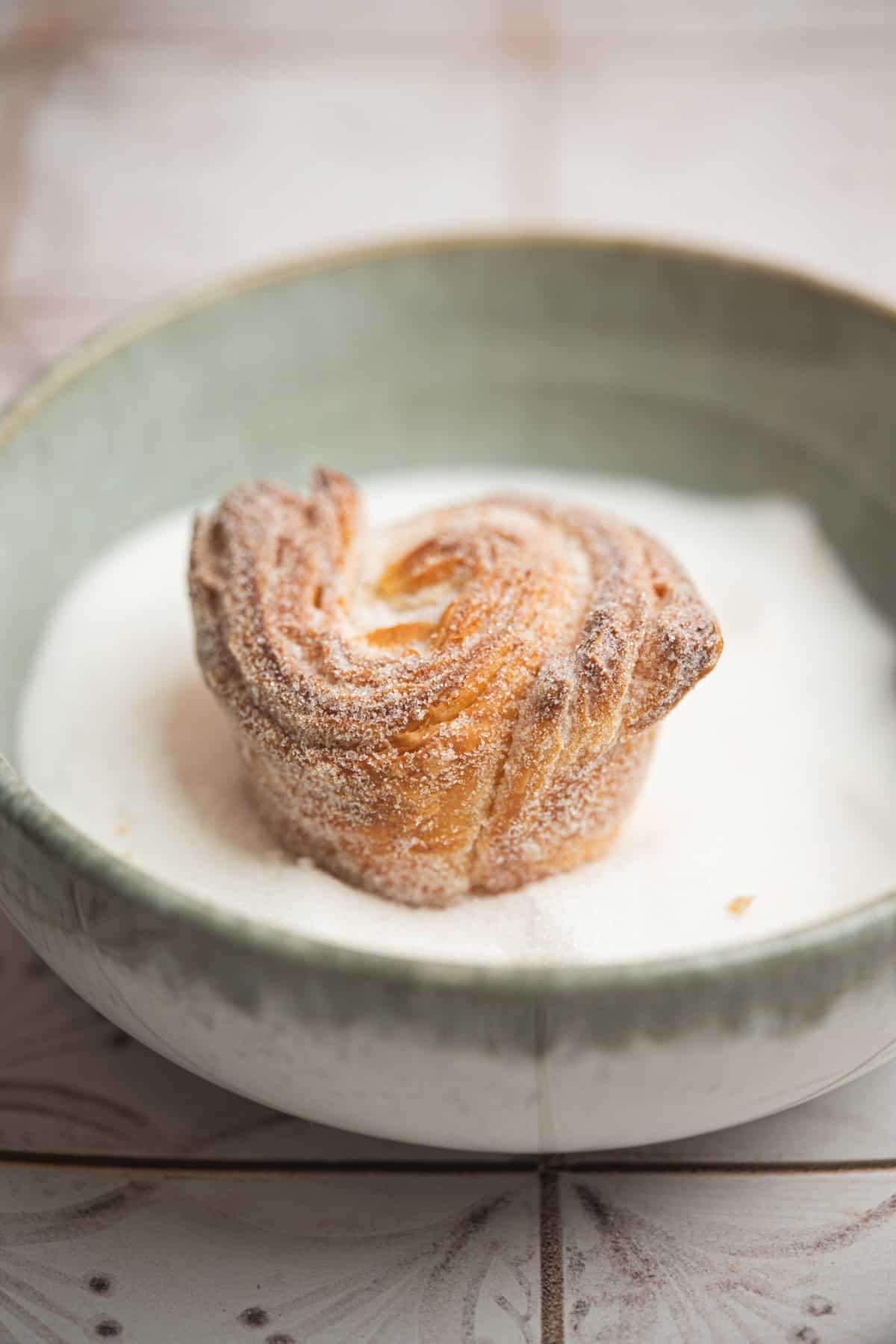 A single sugar-dusted pastry sits in a greenish-gray bowl filled with white sugar, on a light-colored surface. The pastry has a swirled, flaky texture.