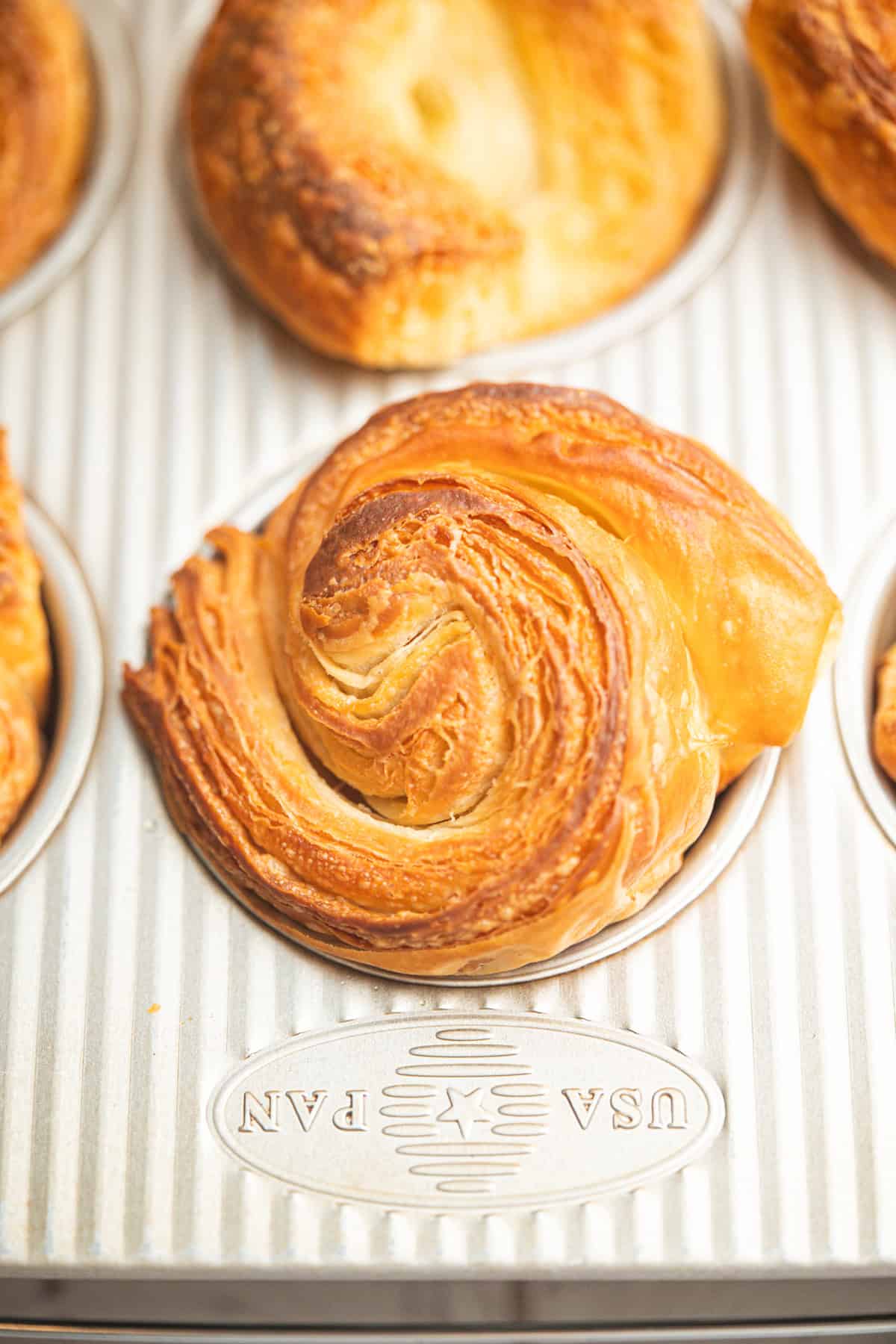 A golden-brown, flaky pastry with swirled layers sits in a metal muffin tin marked with USA PAN. Other pastries are partially visible in the background.