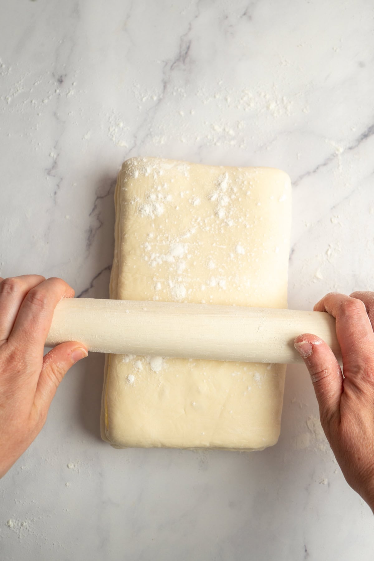 A pair of hands uses a rolling pin to roll out a rectangular block of dough on a lightly floured marble surface.