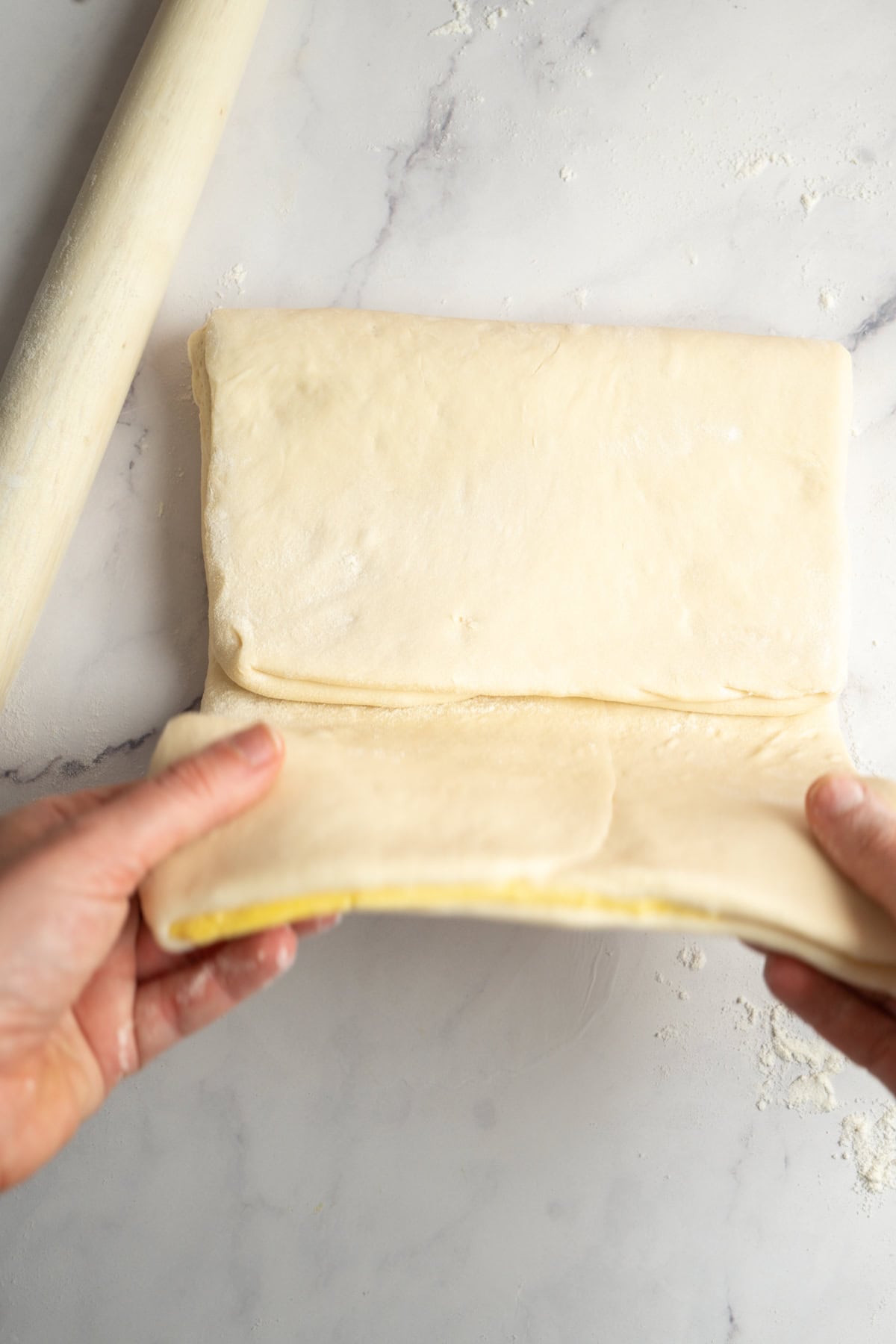 Hands folding a sheet of rolled-out dough on a floured surface, with a rolling pin nearby.