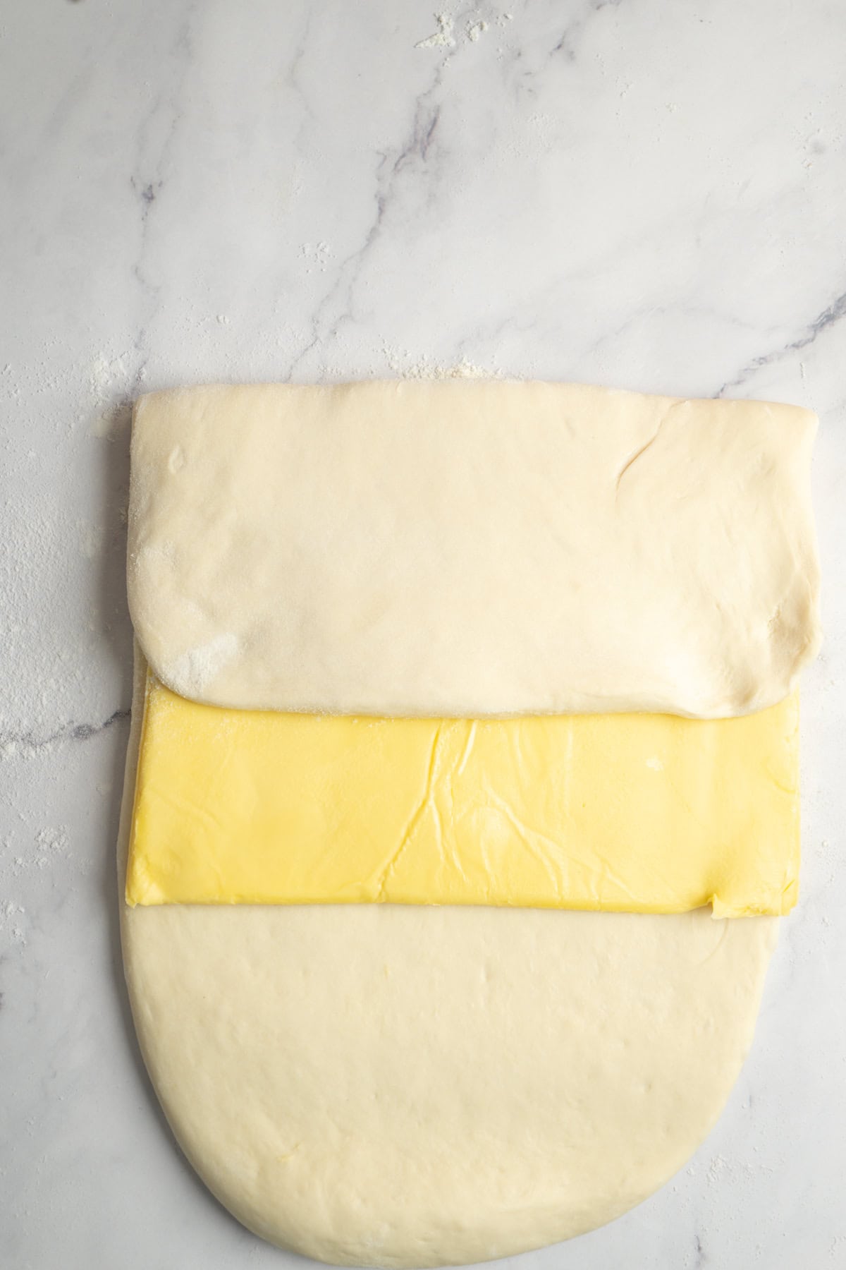 A sheet of dough is laid out on a floured, light marble surface, with a rectangular slab of butter layered on top and the upper portion of dough folded halfway over the butter.