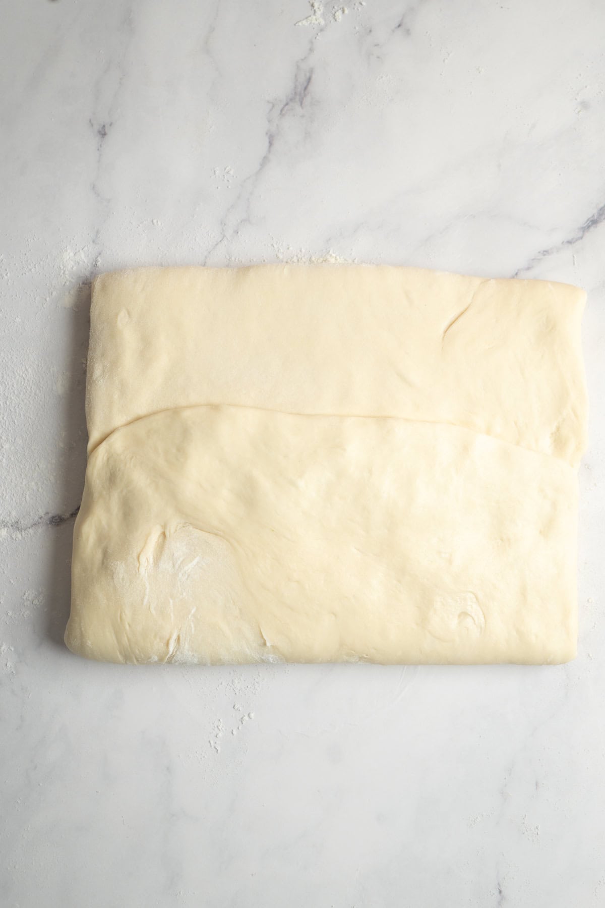 A sheet of dough is folded over itself on a lightly floured marble surface, ready for further shaping or rolling.
