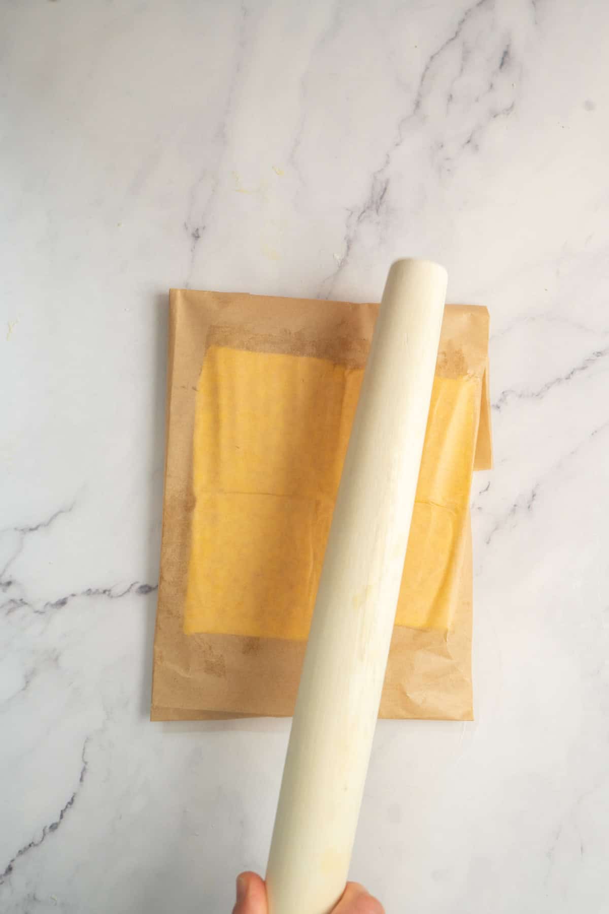 A hand holding a rolling pin, flattening dough between two sheets of parchment paper on a light marble surface.