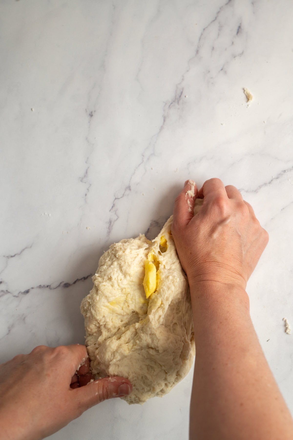 Two hands knead dough with visible chunks of butter on a white marble surface.