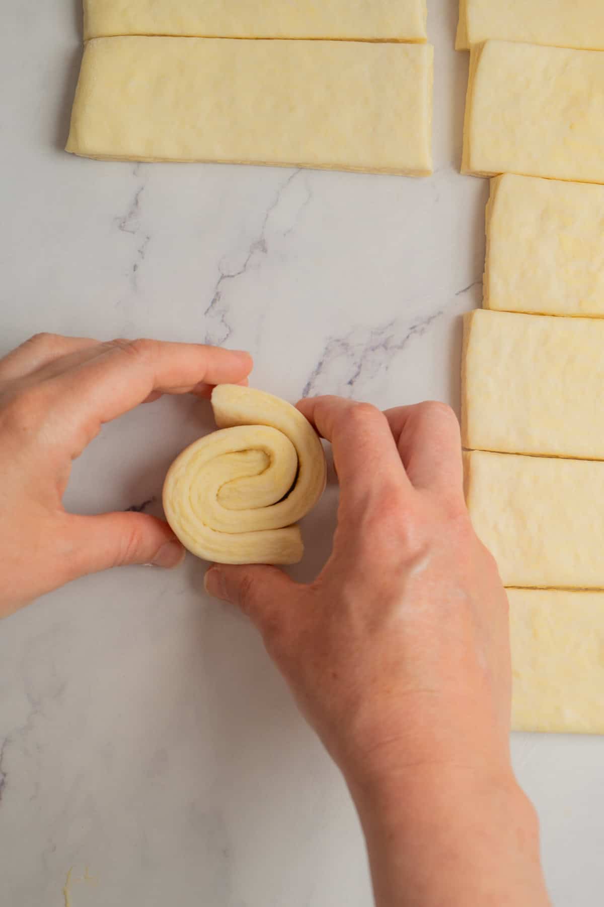 Hands folding a strip of dough into a swirl shape on a marble surface, with several rectangular dough pieces arranged nearby.