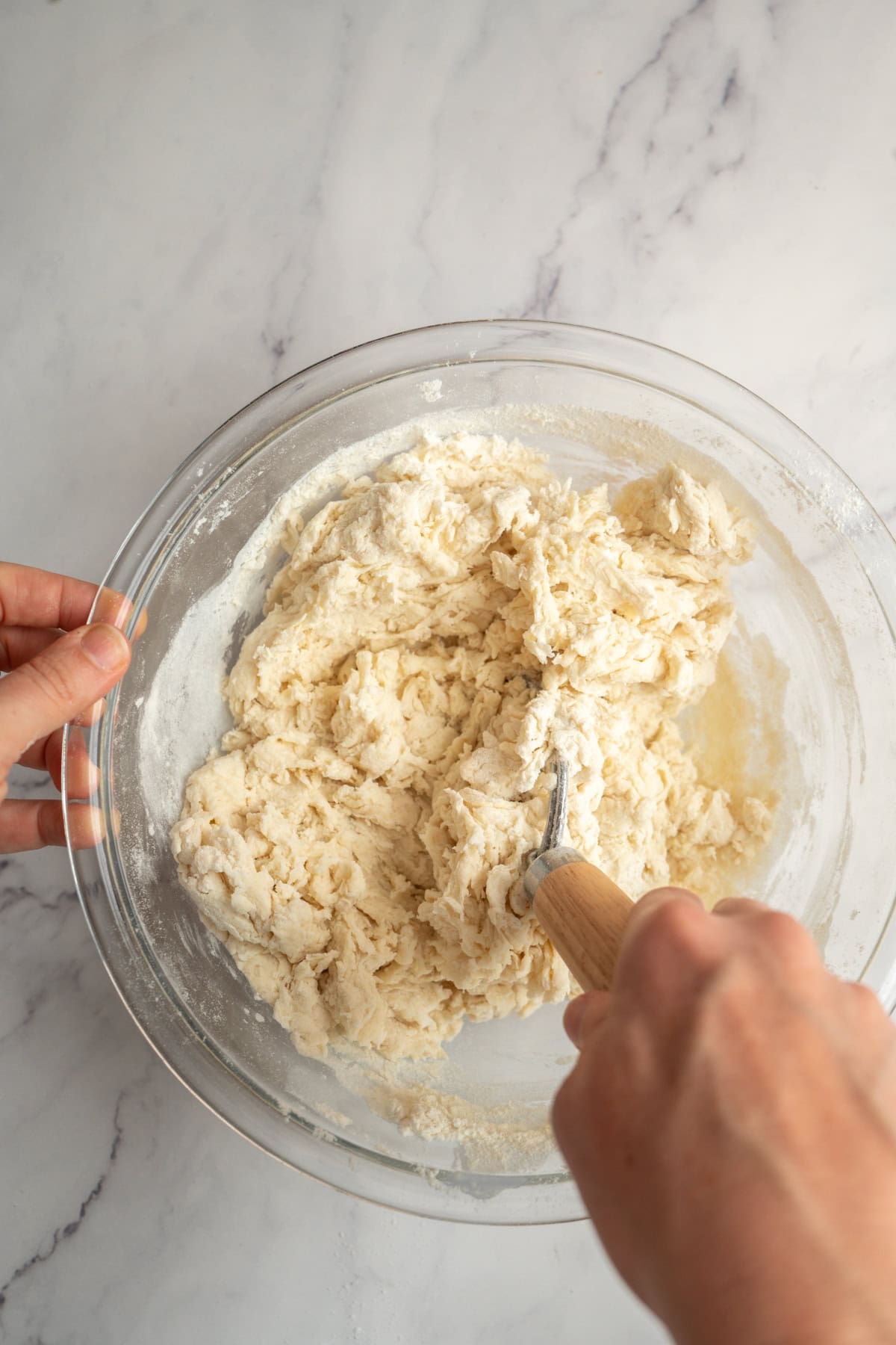 A person’s hands mix flour and liquid ingredients in a large glass bowl using a pastry blender on a white marble countertop.