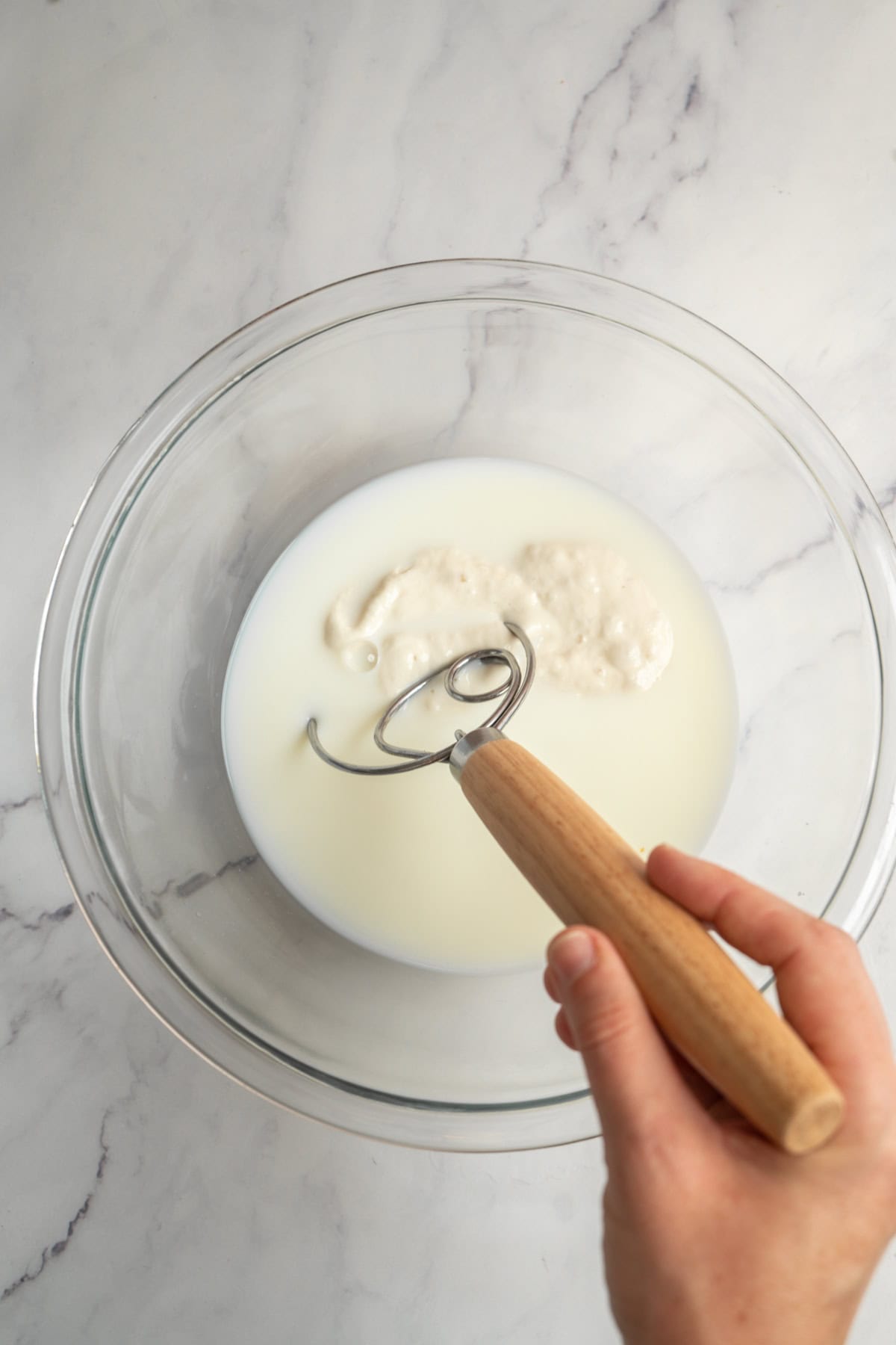 A hand holding a dough whisk stirs sourdough starter and milk in a clear glass bowl on a white marble surface.