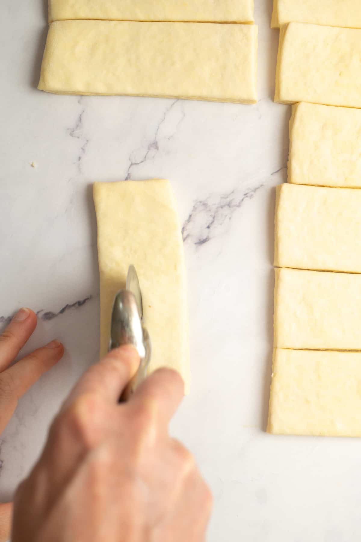 A hand uses a pastry cutter to slice rectangular pieces of dough on a white marble surface. Several evenly cut dough strips are arranged nearby.
