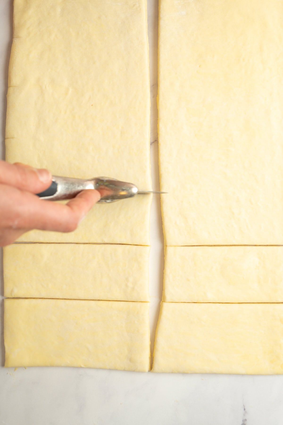 A hand holding a pizza cutter slices rectangular sheets of dough into even strips on a light-colored surface.