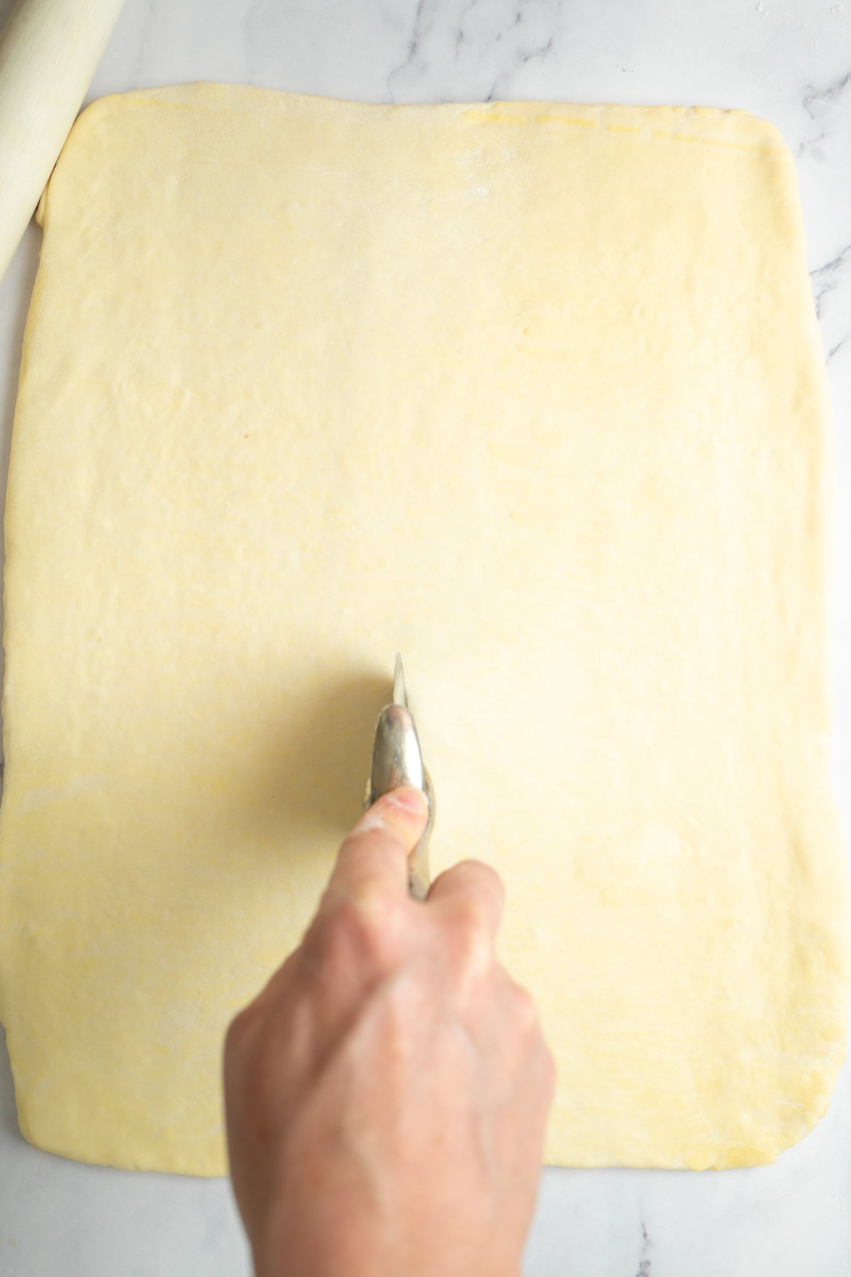 A hand uses a pizza cutter to slice a large, rectangular sheet of dough on a white marble surface.