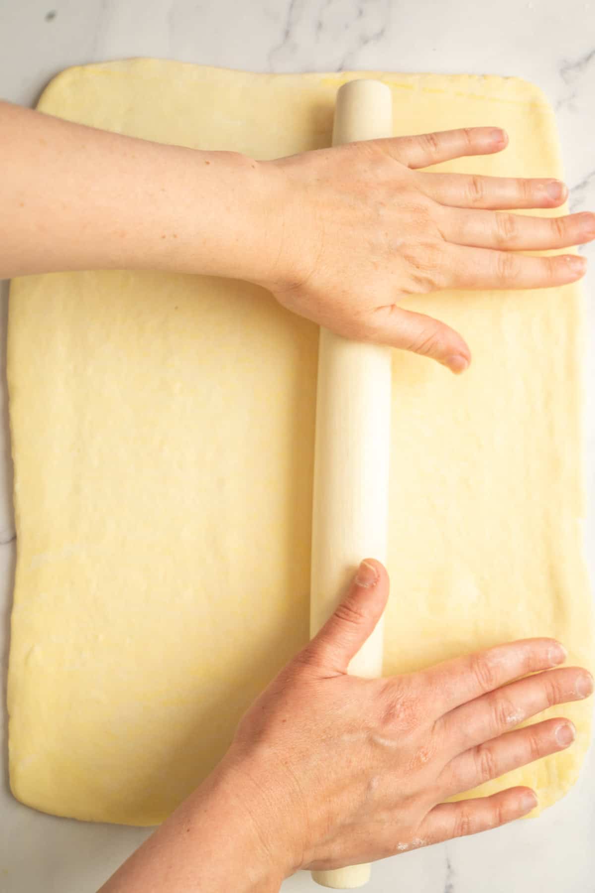 A person uses a rolling pin to flatten a rectangular sheet of dough on a light-colored surface.