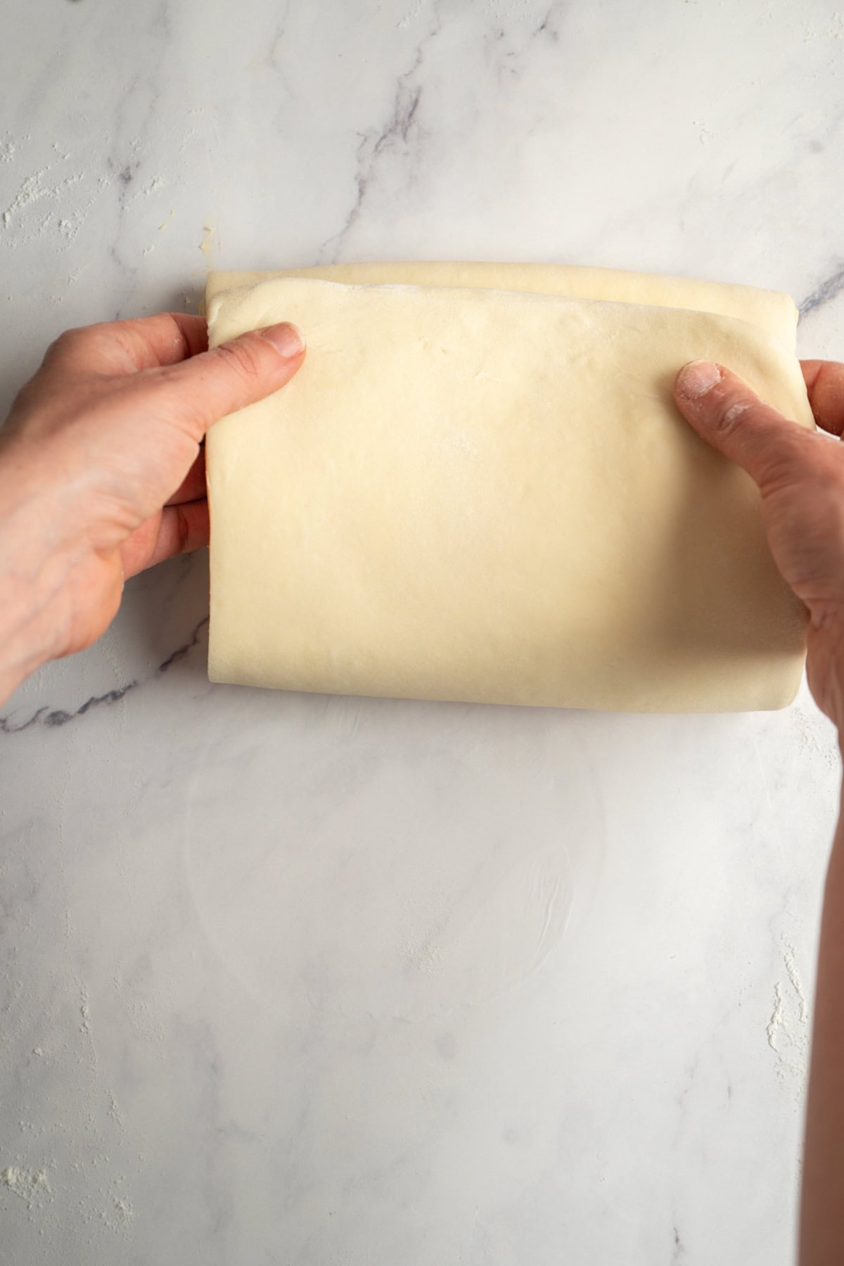Two hands are folding a sheet of pale, smooth dough on a lightly floured, white marble surface.